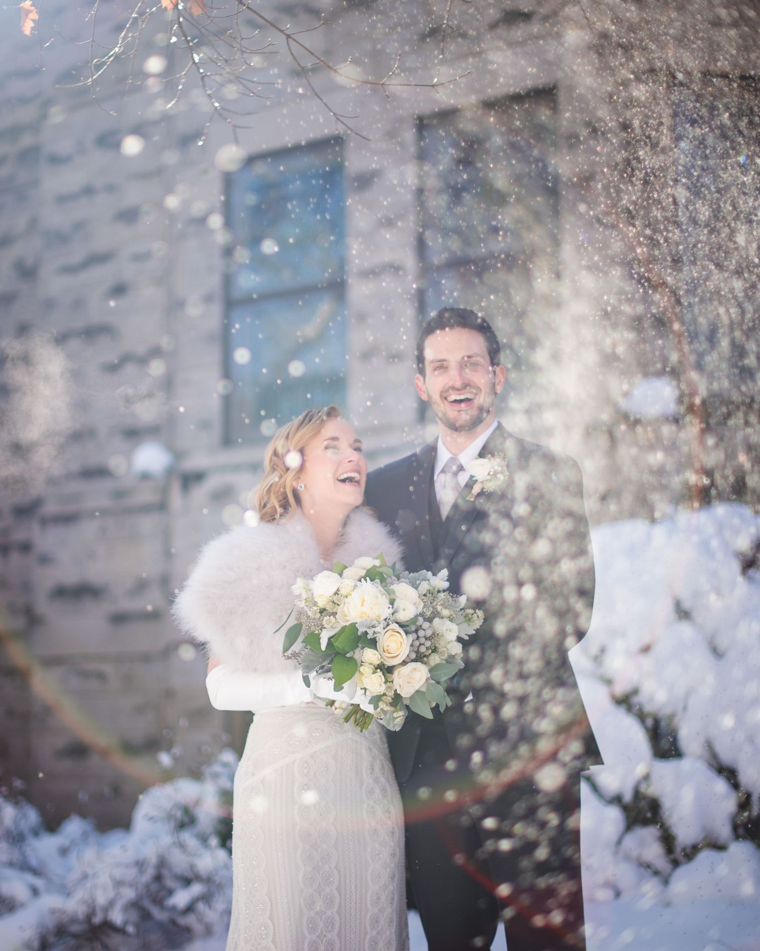 Happy bride and groom laugh together in a magical snowfall outside a stone building in winter.