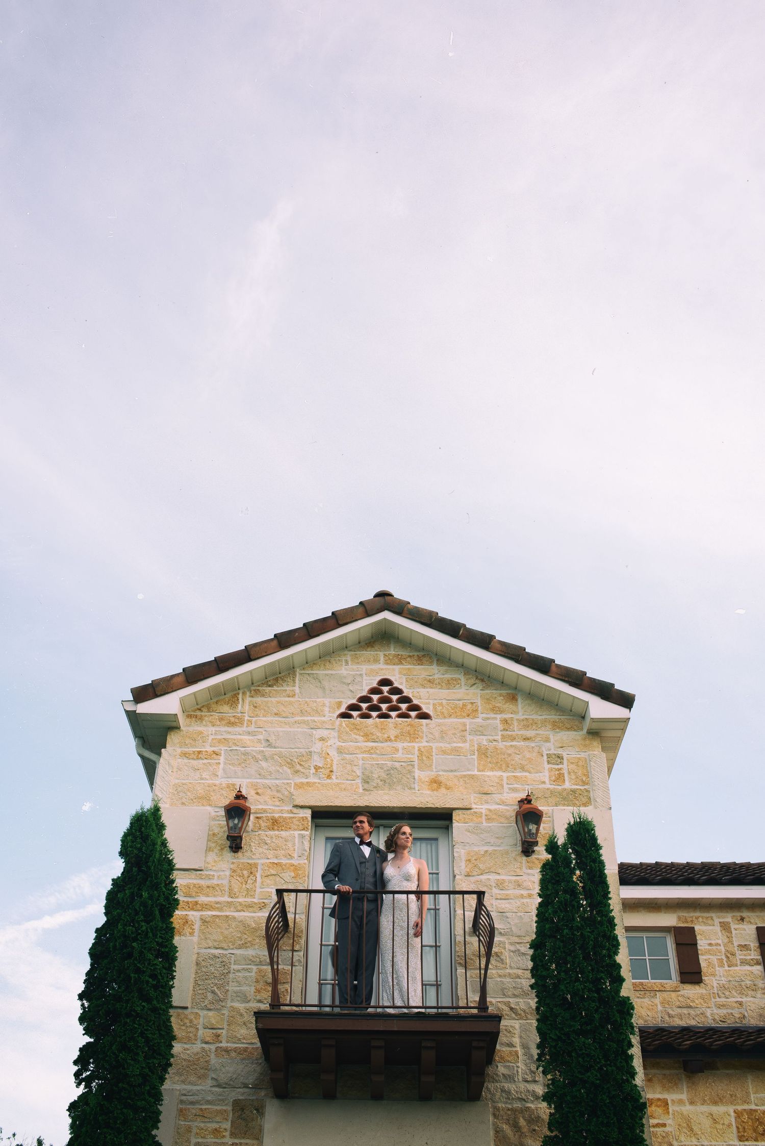 Bride and groom stand together on a stone building balcony under a cloudy sky.