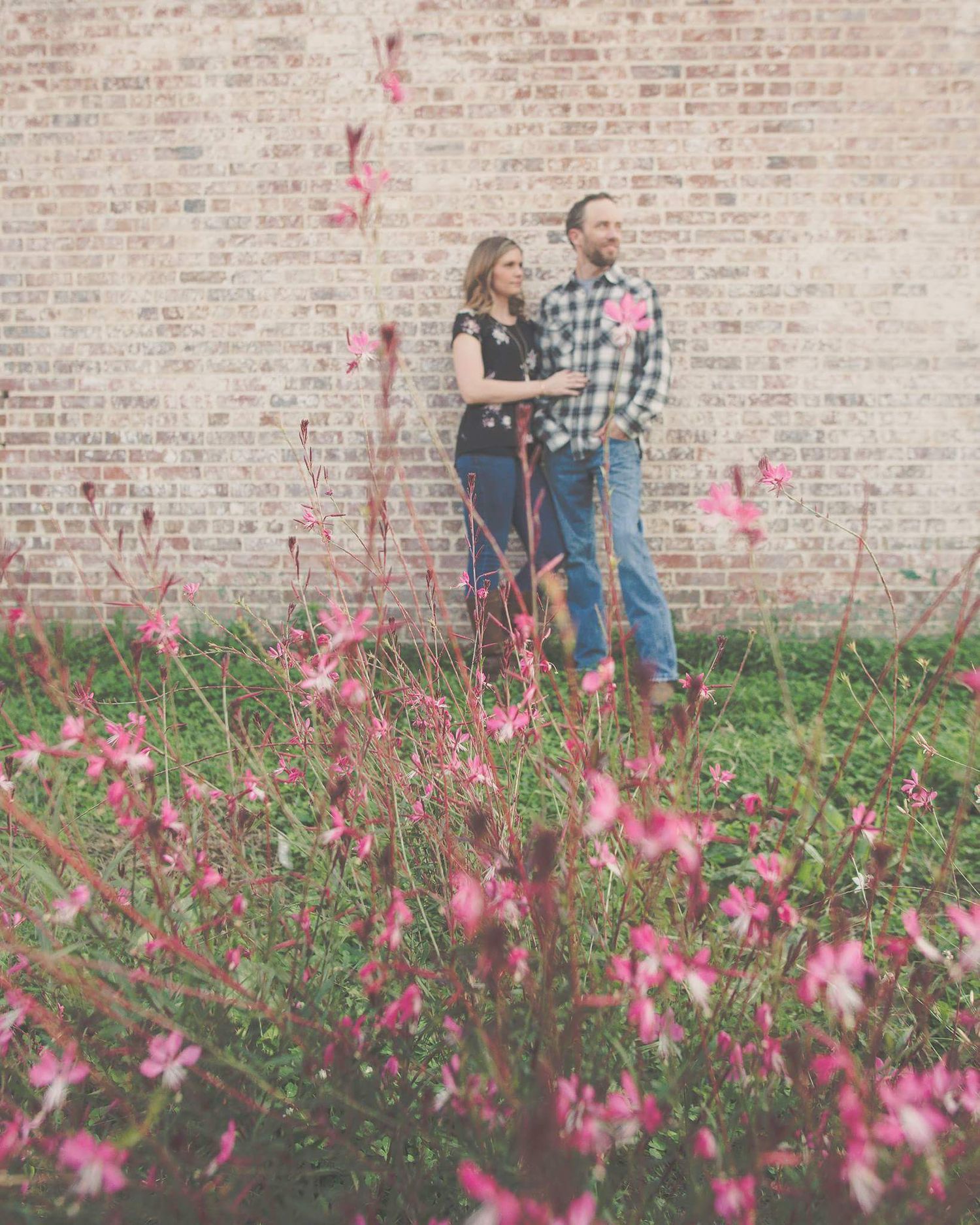 Couple stands together among pink wildflowers against a rustic brick wall during an engagement shoot.