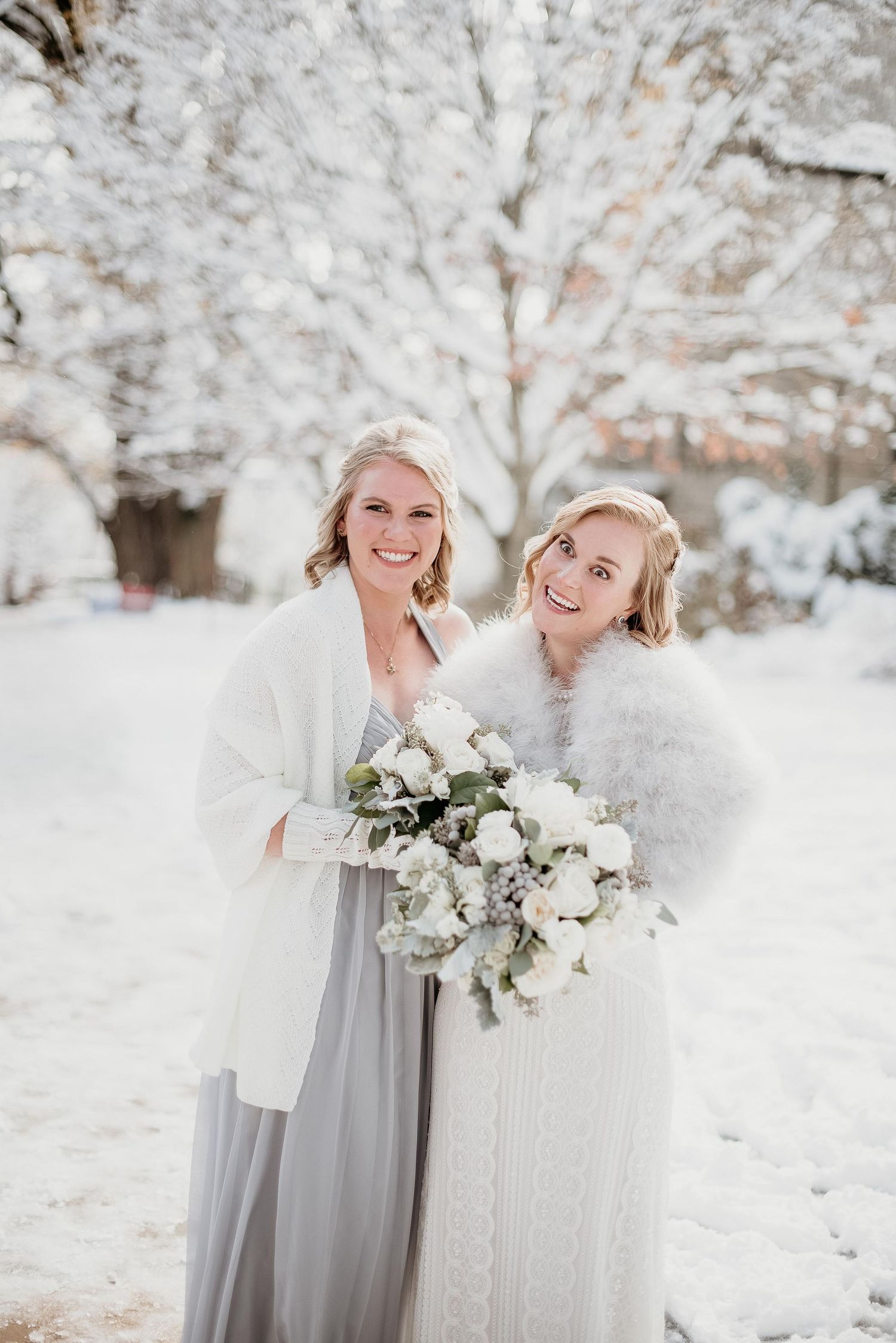 Two brides in white fur wraps hold bouquets of white roses while posing in a snowy winter wonderland setting.