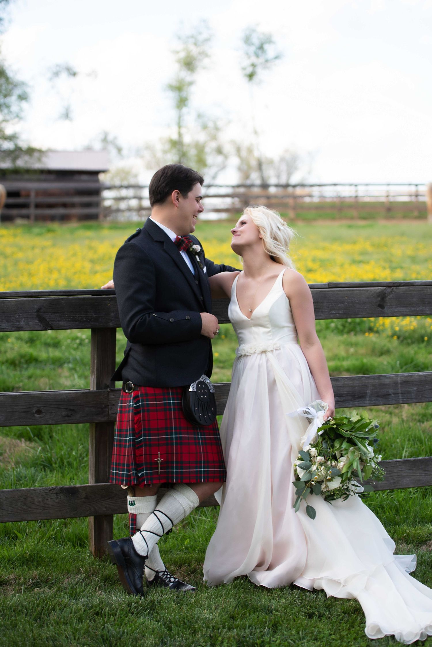Bride in white gown and groom in red tartan kilt share a romantic moment by a rustic wooden fence.