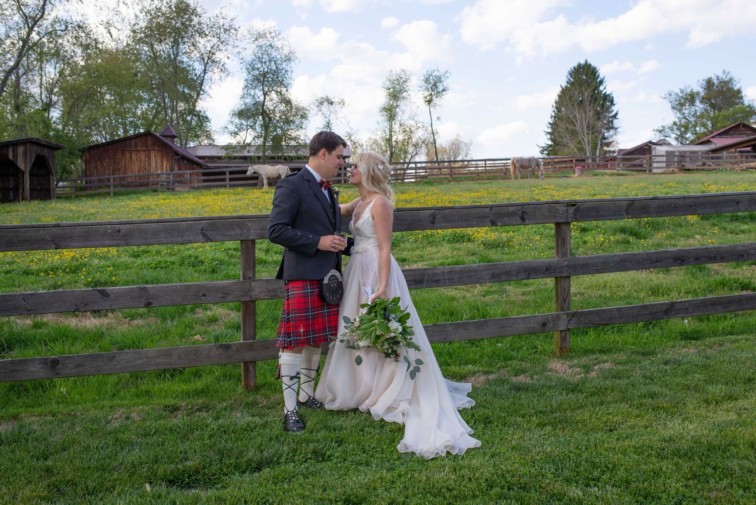 Bride and groom in Scottish kilt pose by rustic wooden fence on green farm grounds.
