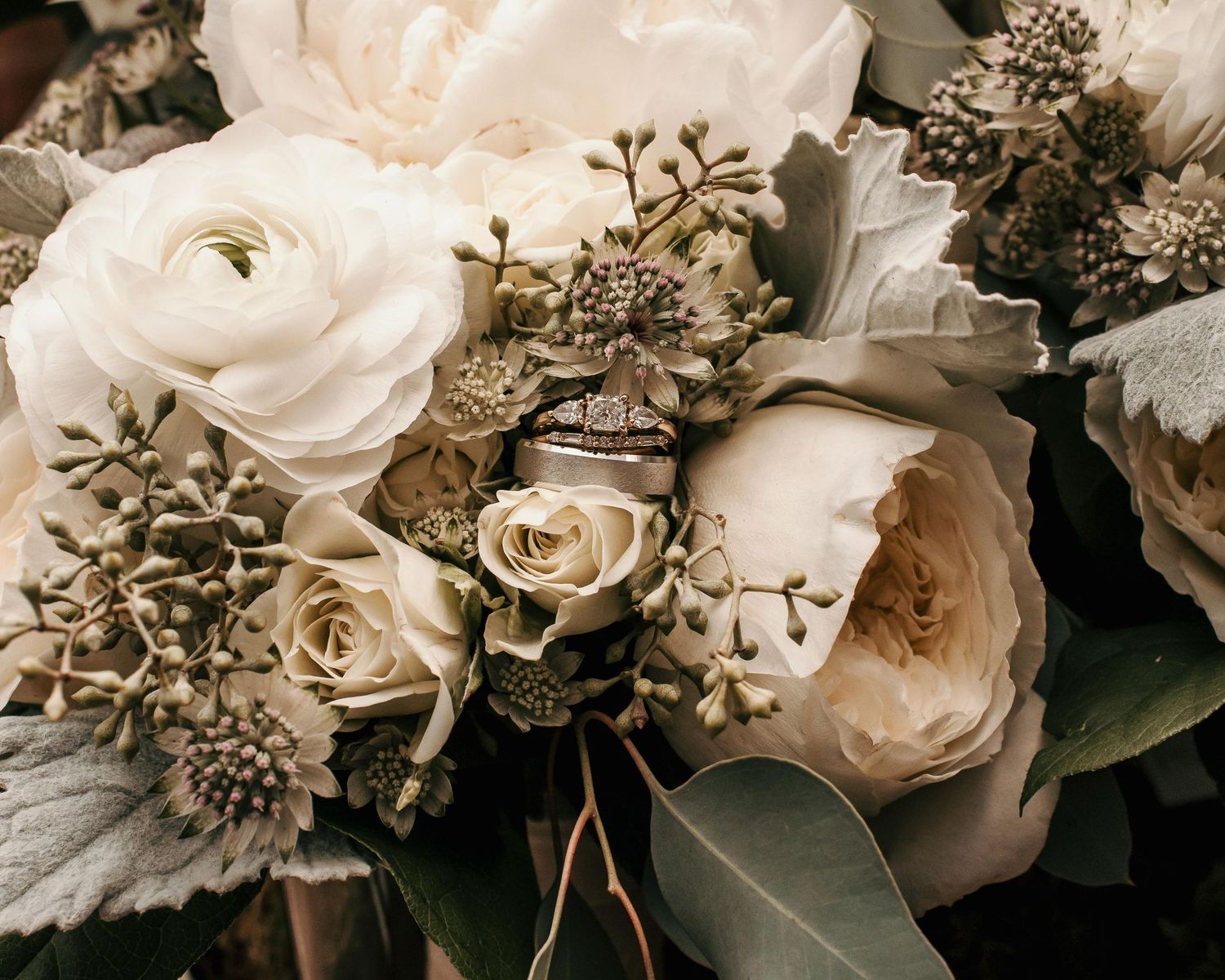 Elegant wedding bouquet with white peonies, roses, and greenery, featuring a sparkling diamond engagement ring nestled among the blooms.