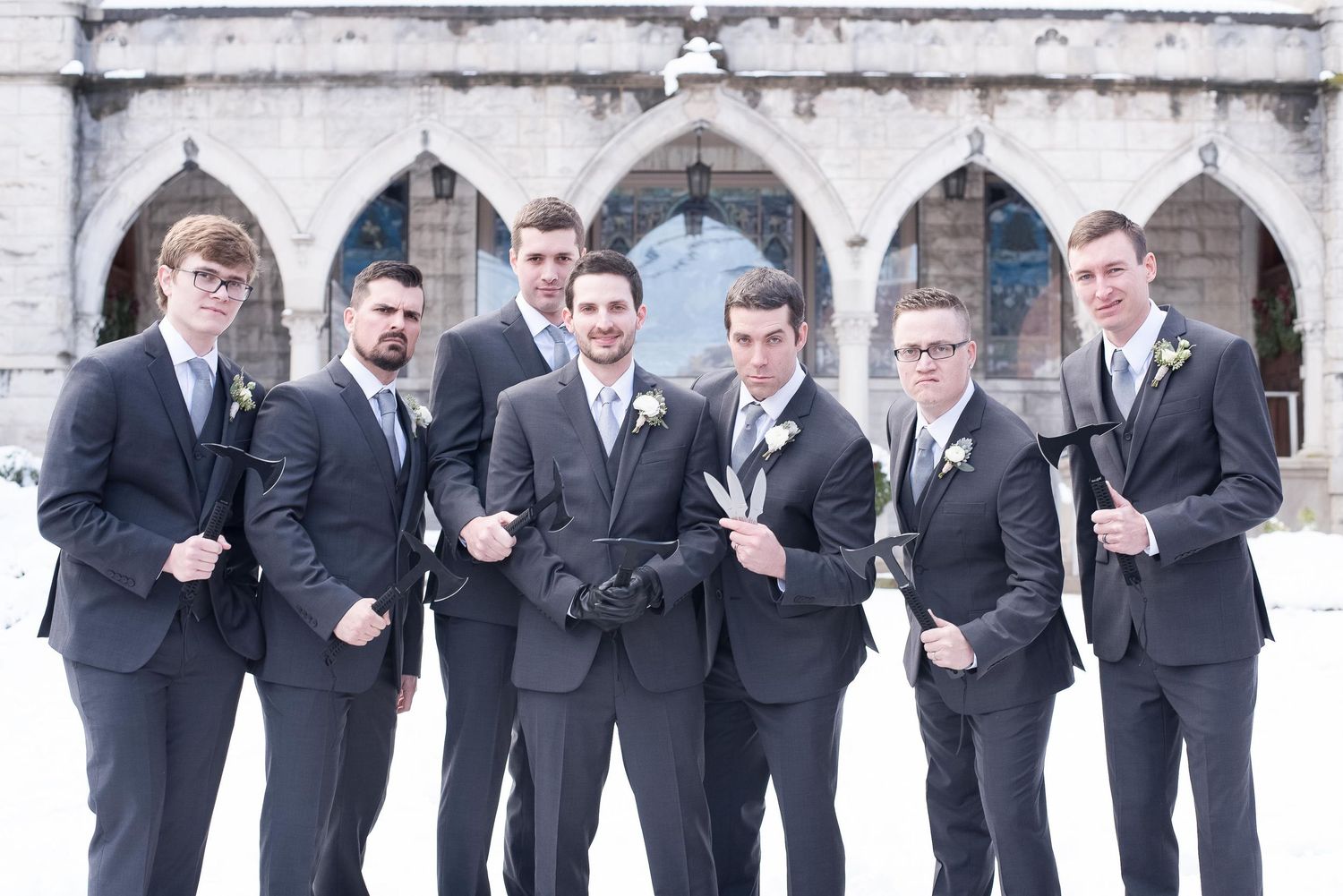 Seven groomsmen in dark suits pose outdoors in snow before a stone church for a winter wedding photo.