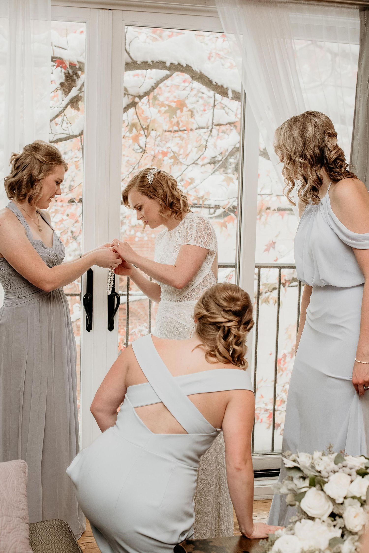 Bridesmaids in silver gowns help bride with final wedding preparations near a bright window with floral views.