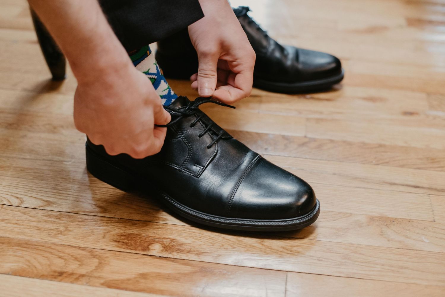 Man tying black leather dress shoes on hardwood floor, wearing dark suit pants and colorful patterned socks.