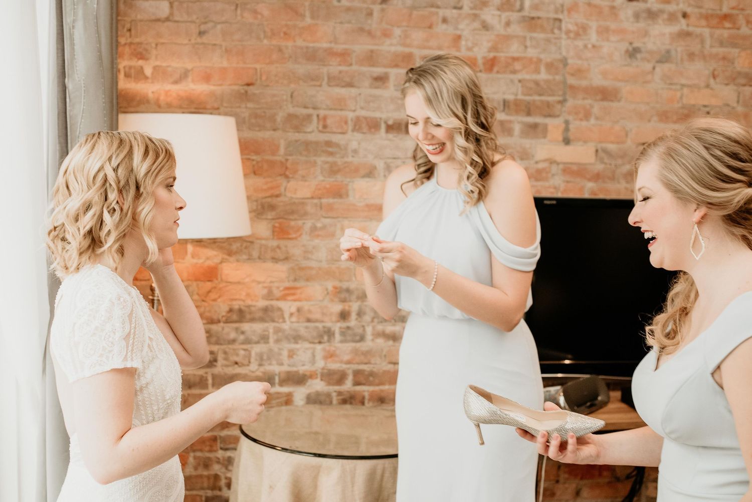 Three bridesmaids in white dresses share a joyful moment in a rustic brick-walled room before a wedding ceremony.