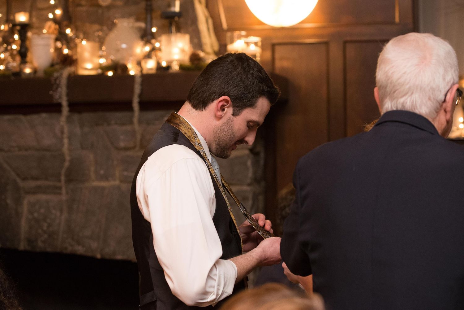 Man in vest and tie adjusting clothing near a fireplace at an elegant indoor event.