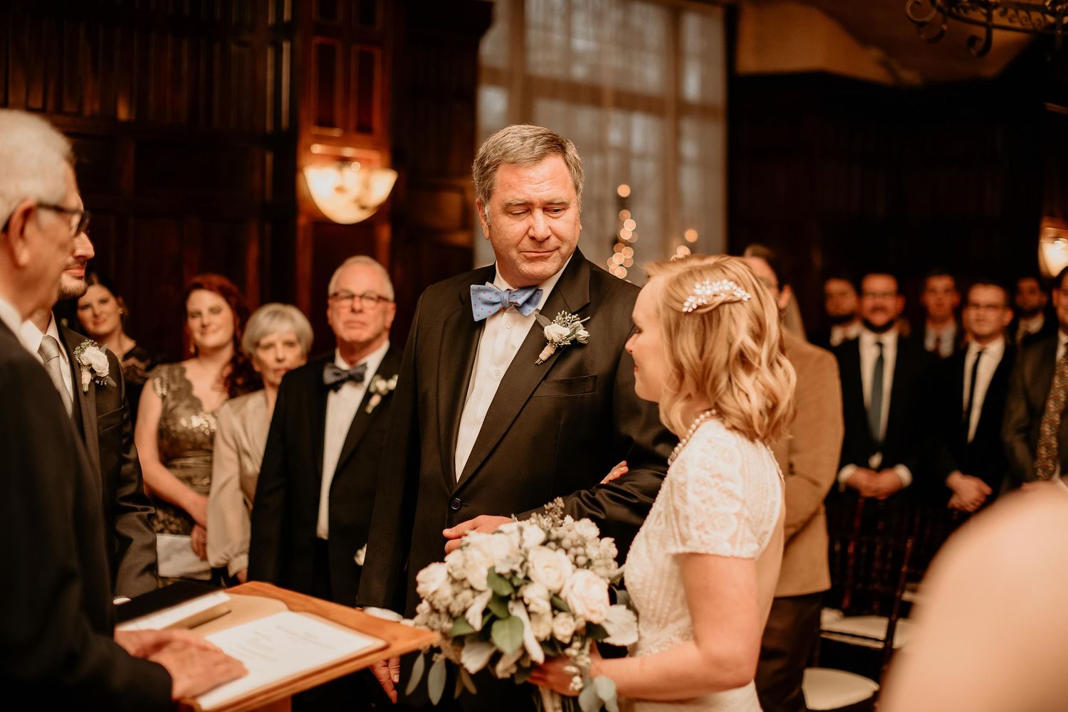 Bride and groom exchanging vows at elegant indoor wedding ceremony with guests standing in background.