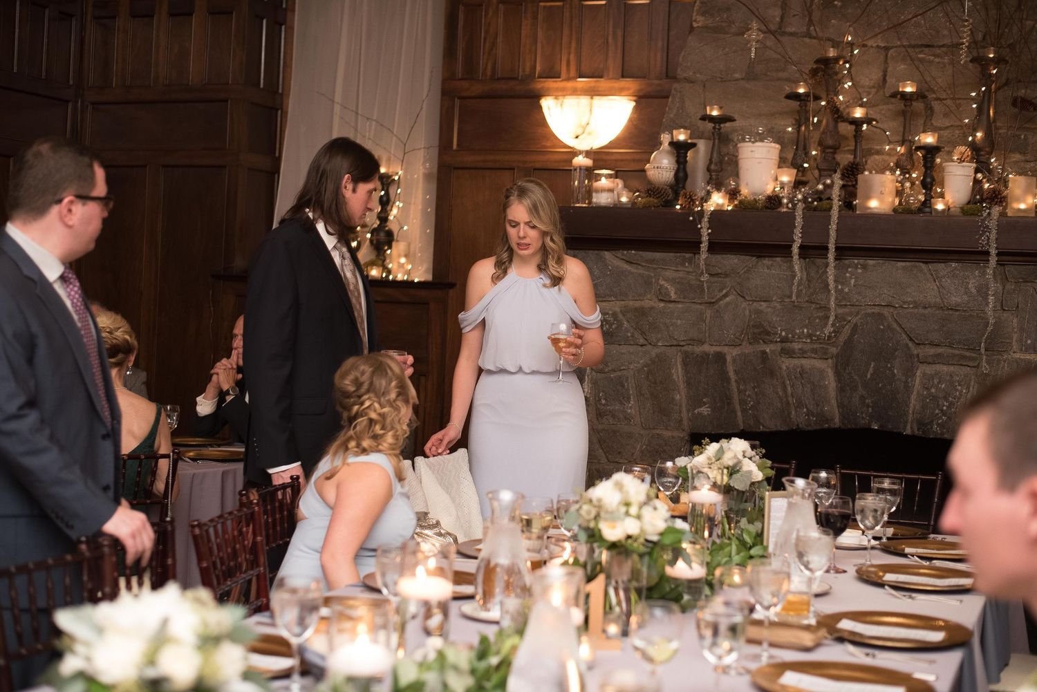 Elegant wedding reception with guests gathered around candlelit tables near a stone fireplace in a rustic venue.