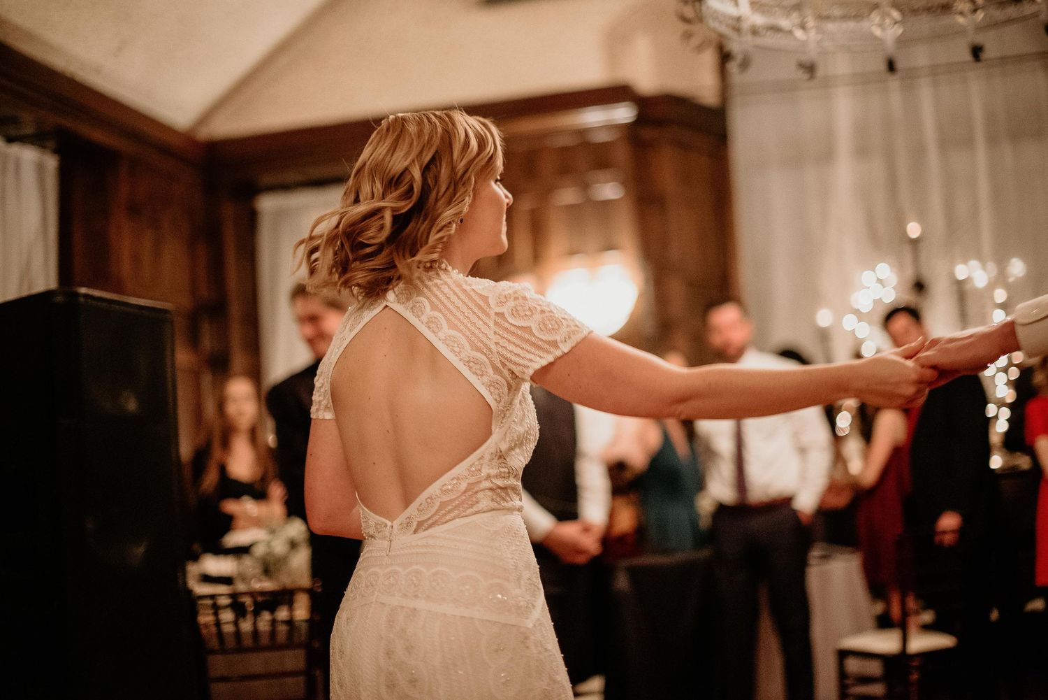 Bride in lace open-back wedding dress dancing at elegant indoor wedding reception with guests in background.