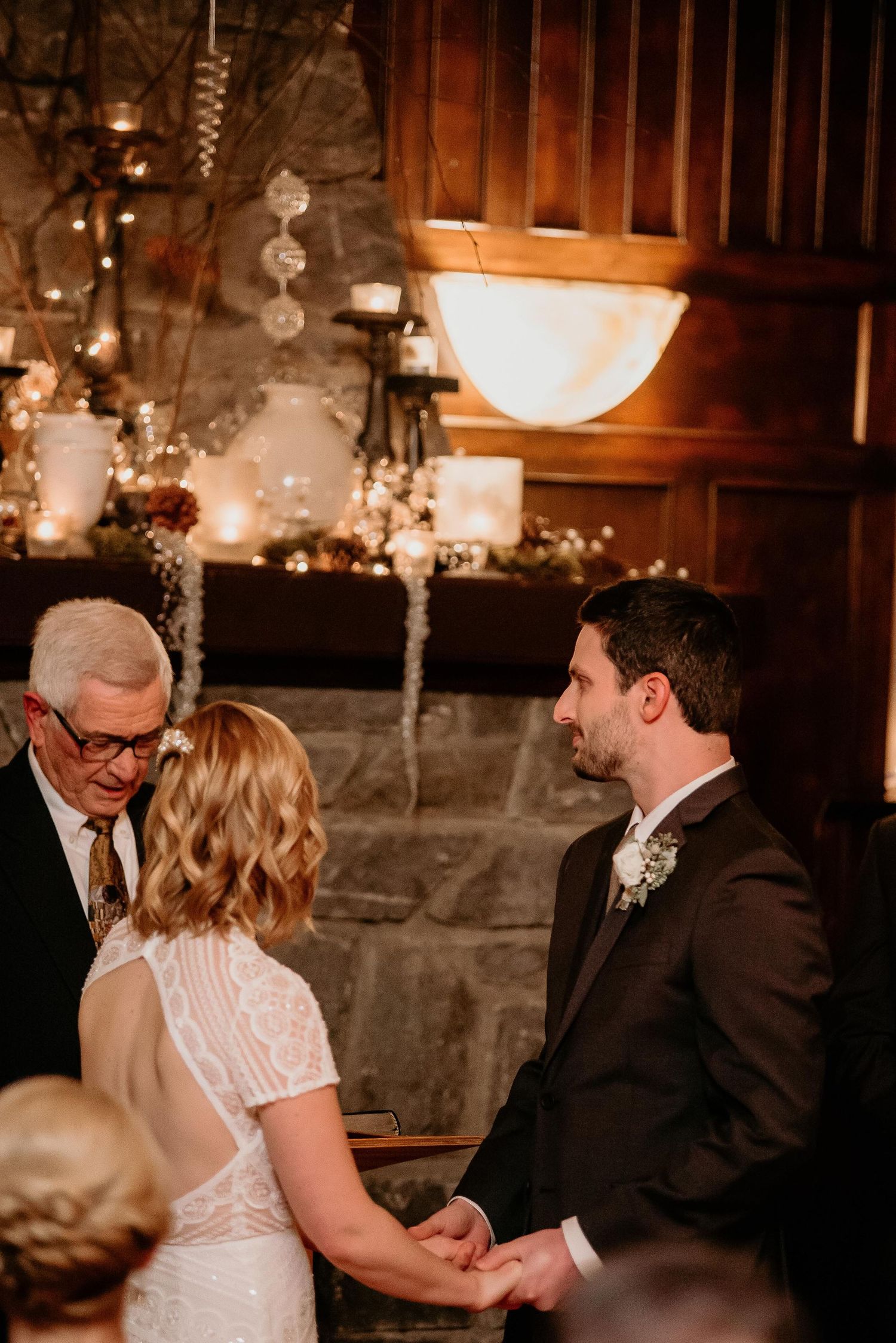 Bride and groom hold hands during intimate indoor wedding ceremony beside stone fireplace with warm candlelight.