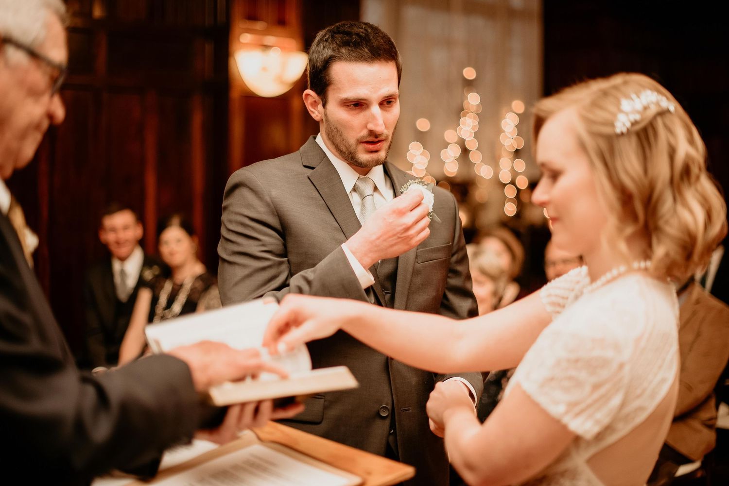 Groom reciting wedding vows to bride during indoor ceremony, with officiant holding book nearby.