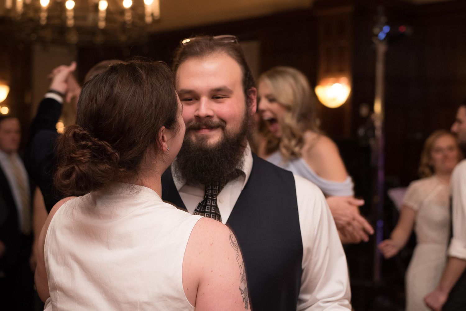 Bearded man in vest and tie smiling while dancing at a wedding reception with guests in background.