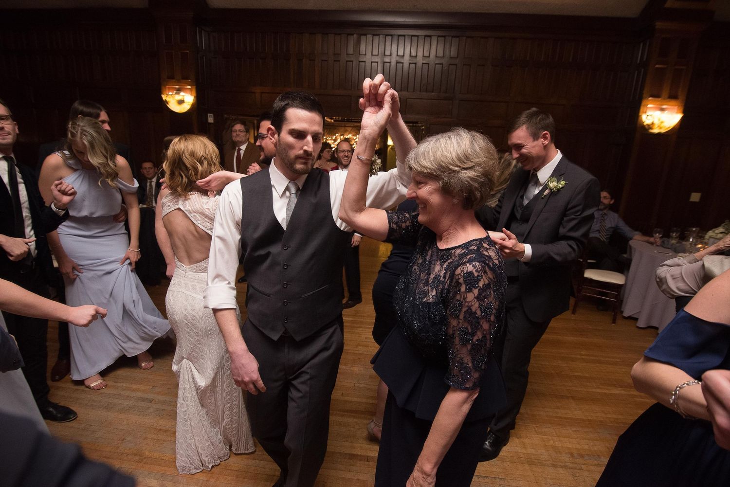 Groom dancing joyfully with older woman at lively wedding reception celebration.
