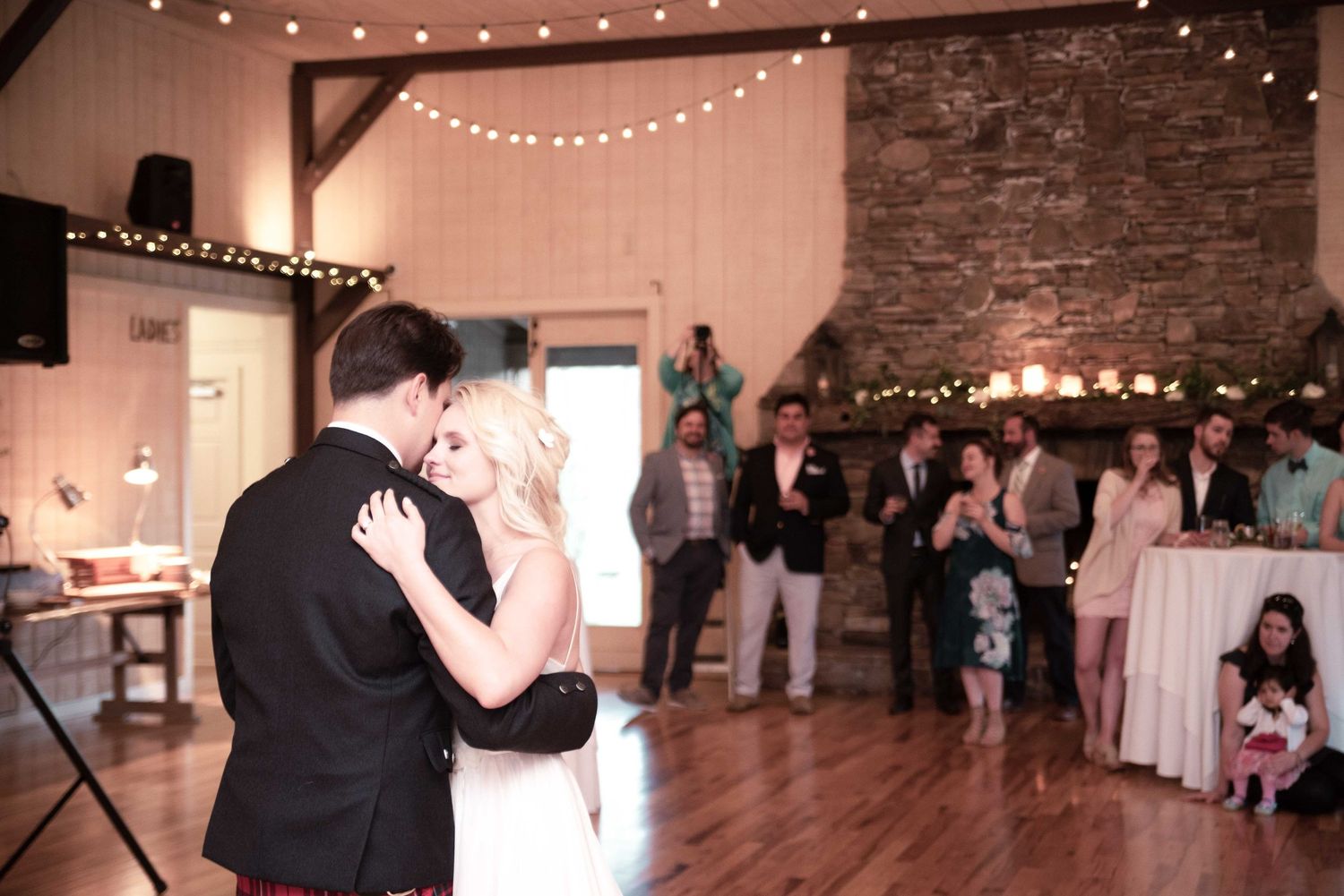 Couple shares first dance at rustic barn wedding reception with string lights and guests watching in background.