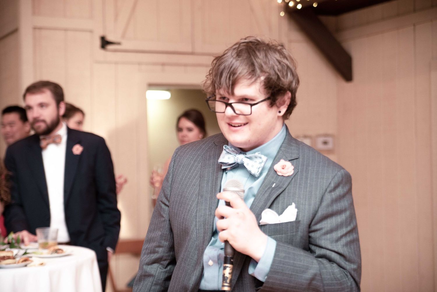 Person in gray suit with bow tie speaks into microphone at formal indoor event.