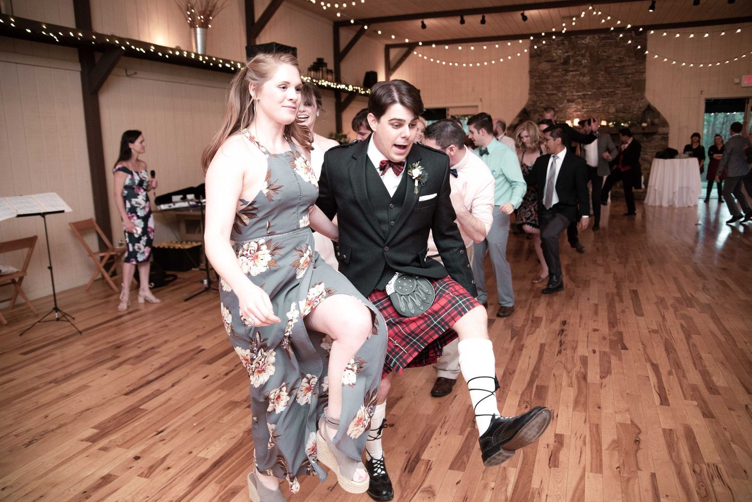 Guests energetically dance at a wedding reception in a rustic venue, with one man wearing a festive red tartan kilt.