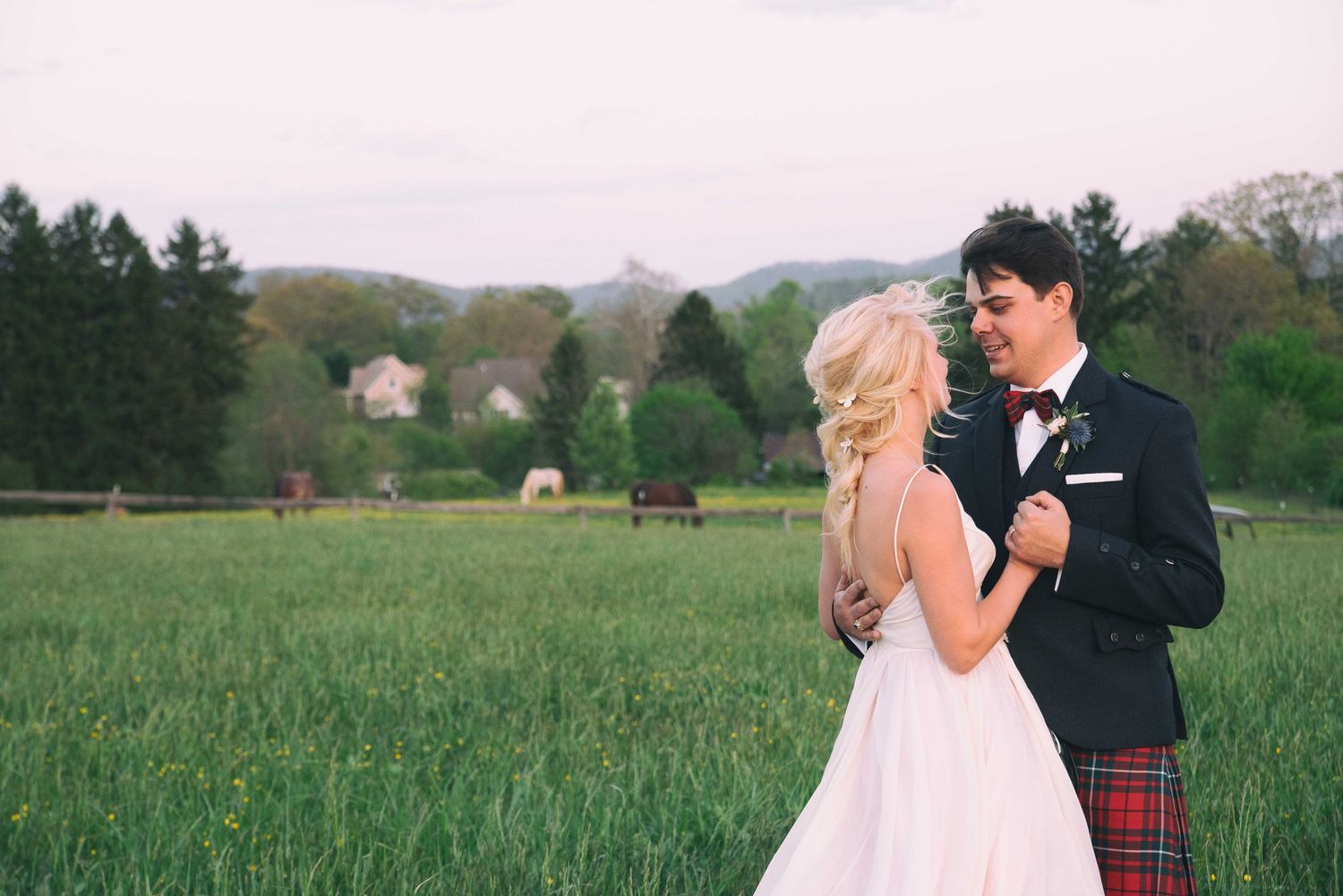 Bride and groom share a romantic dance in a lush green field with horses grazing and rolling hills in the background.