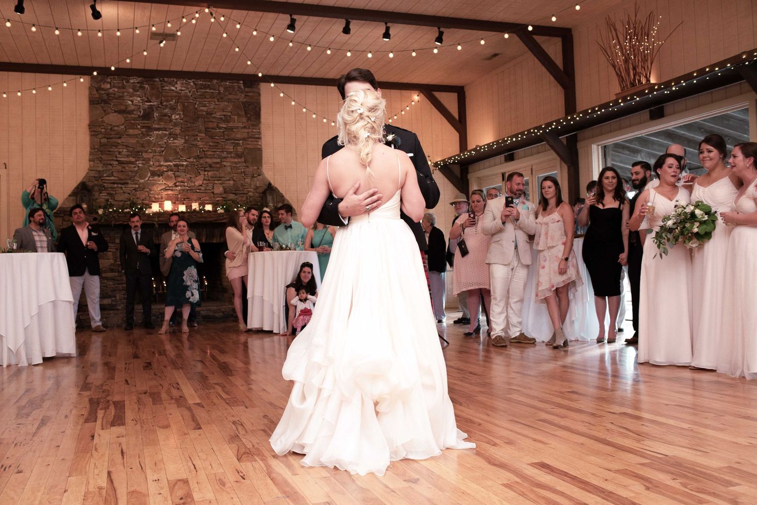 Bride and groom share their first dance on a wooden floor at an elegant rustic wedding venue with guests watching.