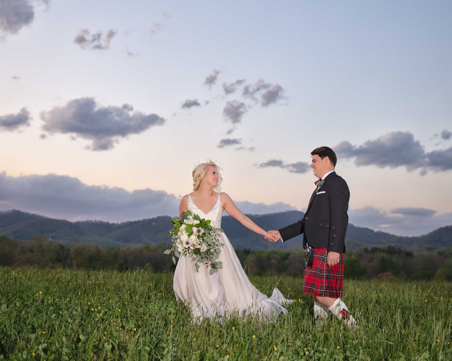 Bride and groom holding hands in green meadow at sunset, groom wearing red Scottish kilt, mountains in background.