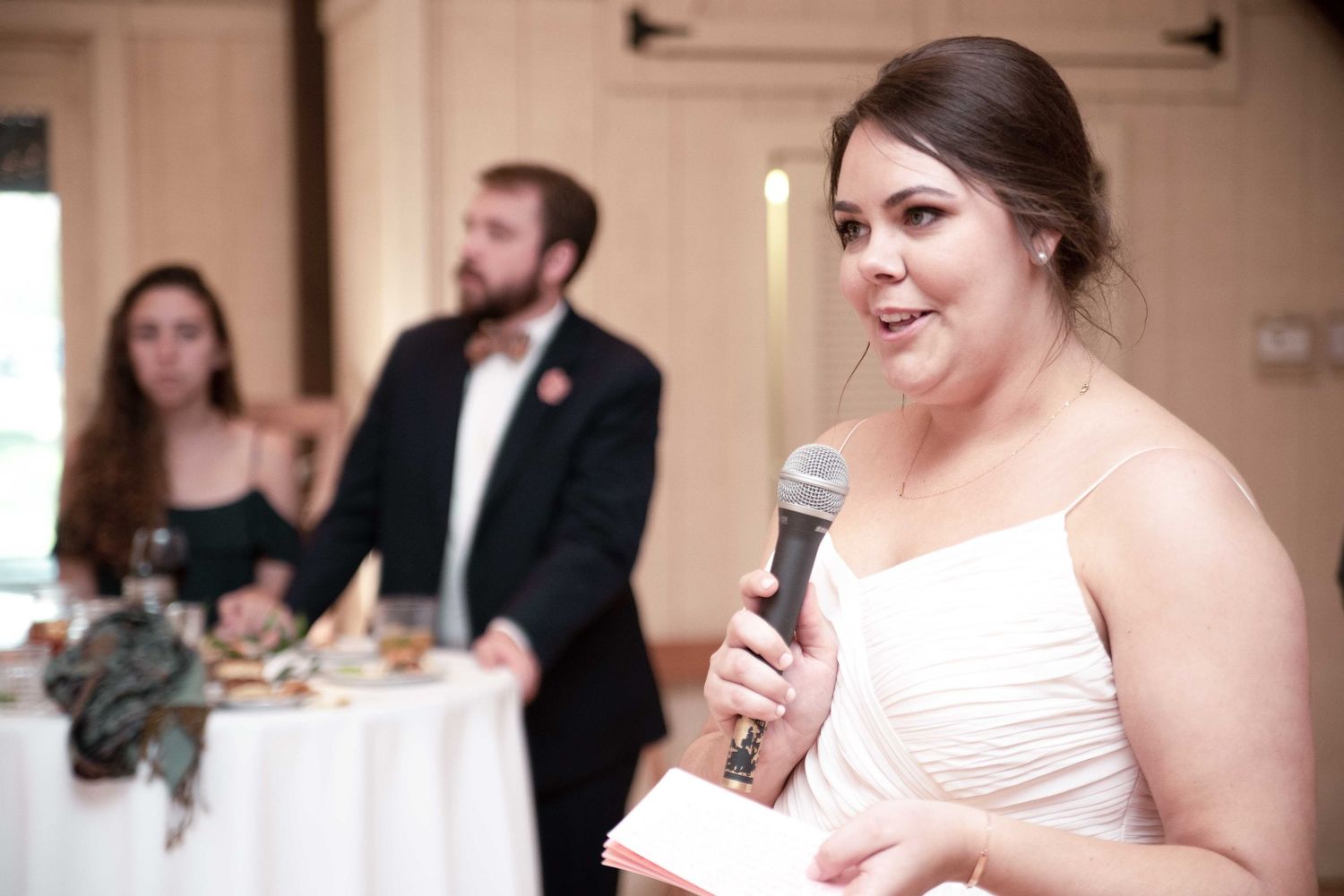 Woman in white dress giving a speech at a wedding reception while holding a microphone and reading from notes.