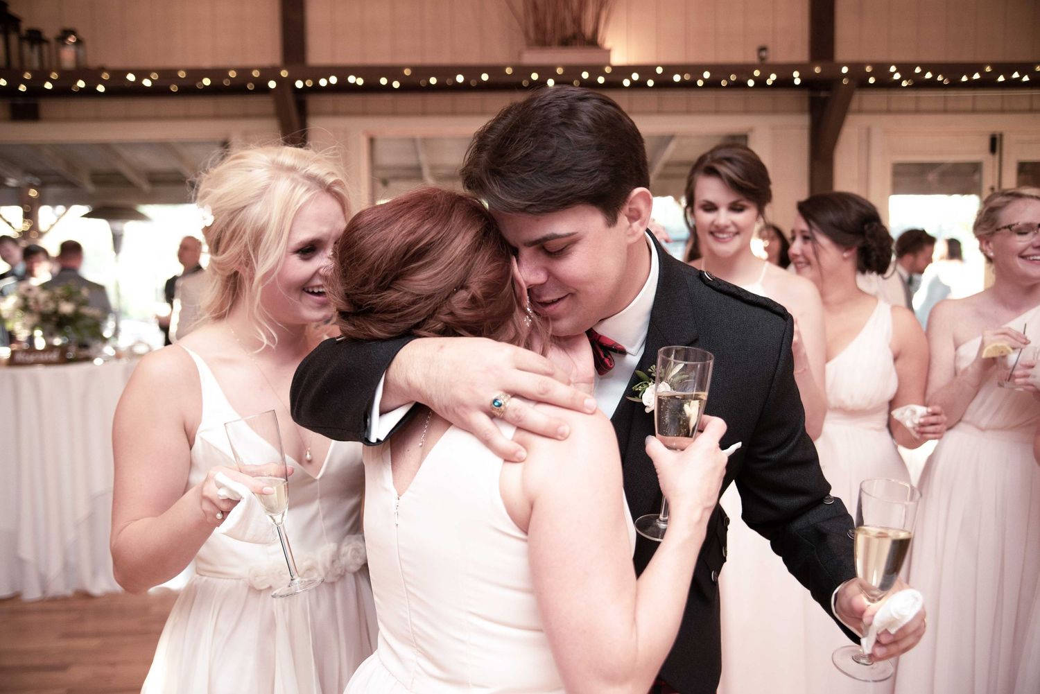 Bride and groom share a warm hug with a bridesmaid during wedding reception, guests celebrating in background.