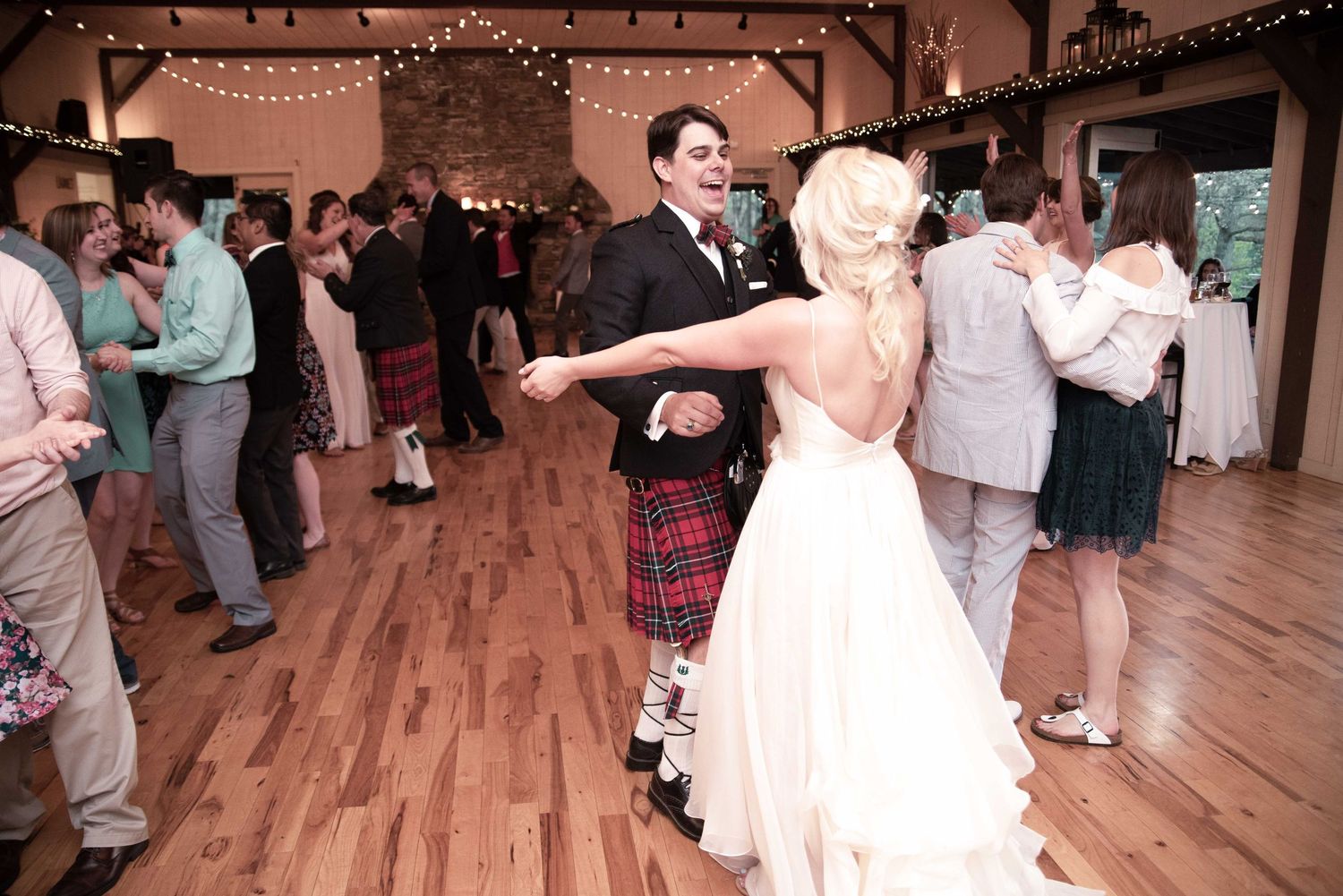 Bride and groom share first dance at wedding reception, groom wearing traditional red tartan kilt in rustic barn venue.