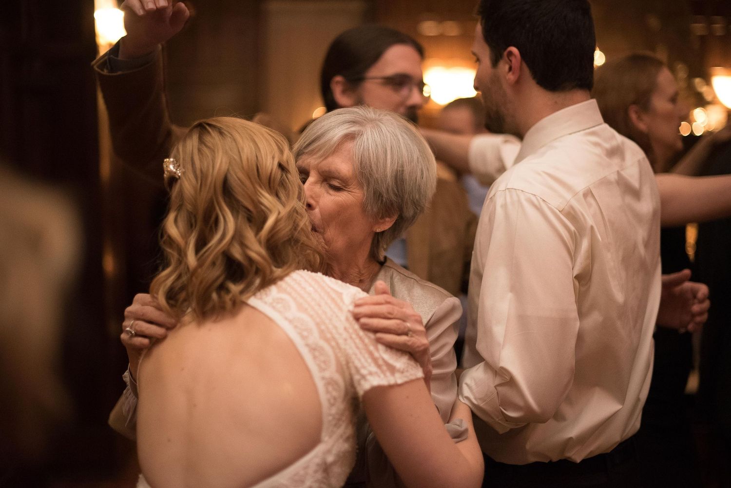 Elderly woman sharing a tender kiss with a bride in a white lace dress at a wedding reception dance floor.