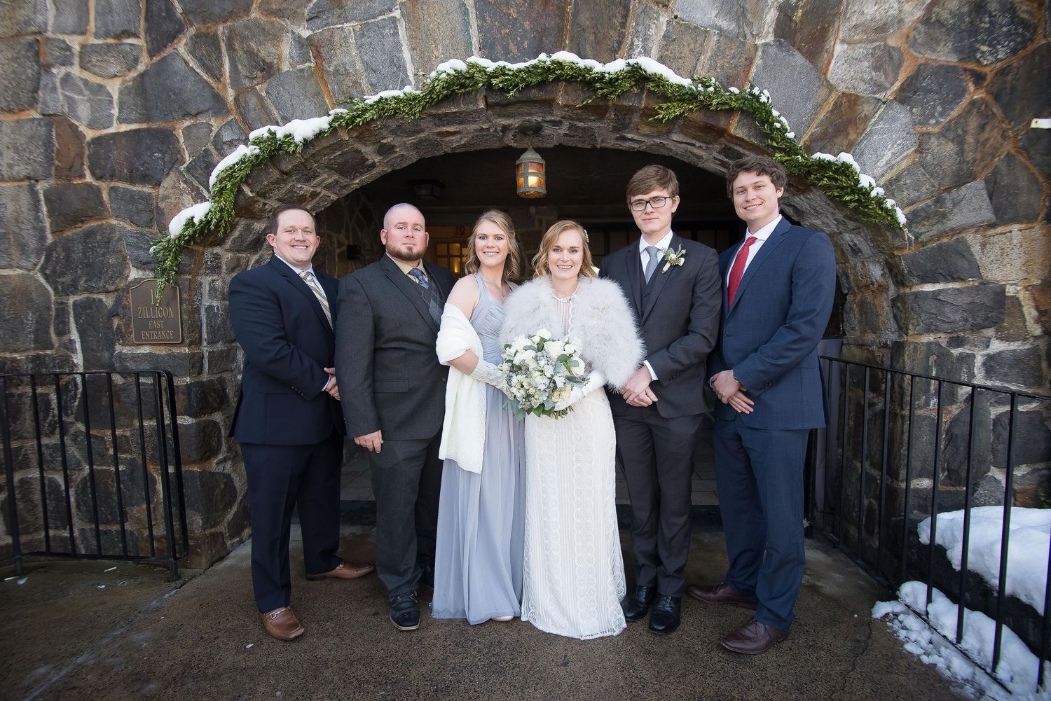 Wedding party posing in formal attire before a stone arch doorway dusted with snow in a winter outdoor setting.
