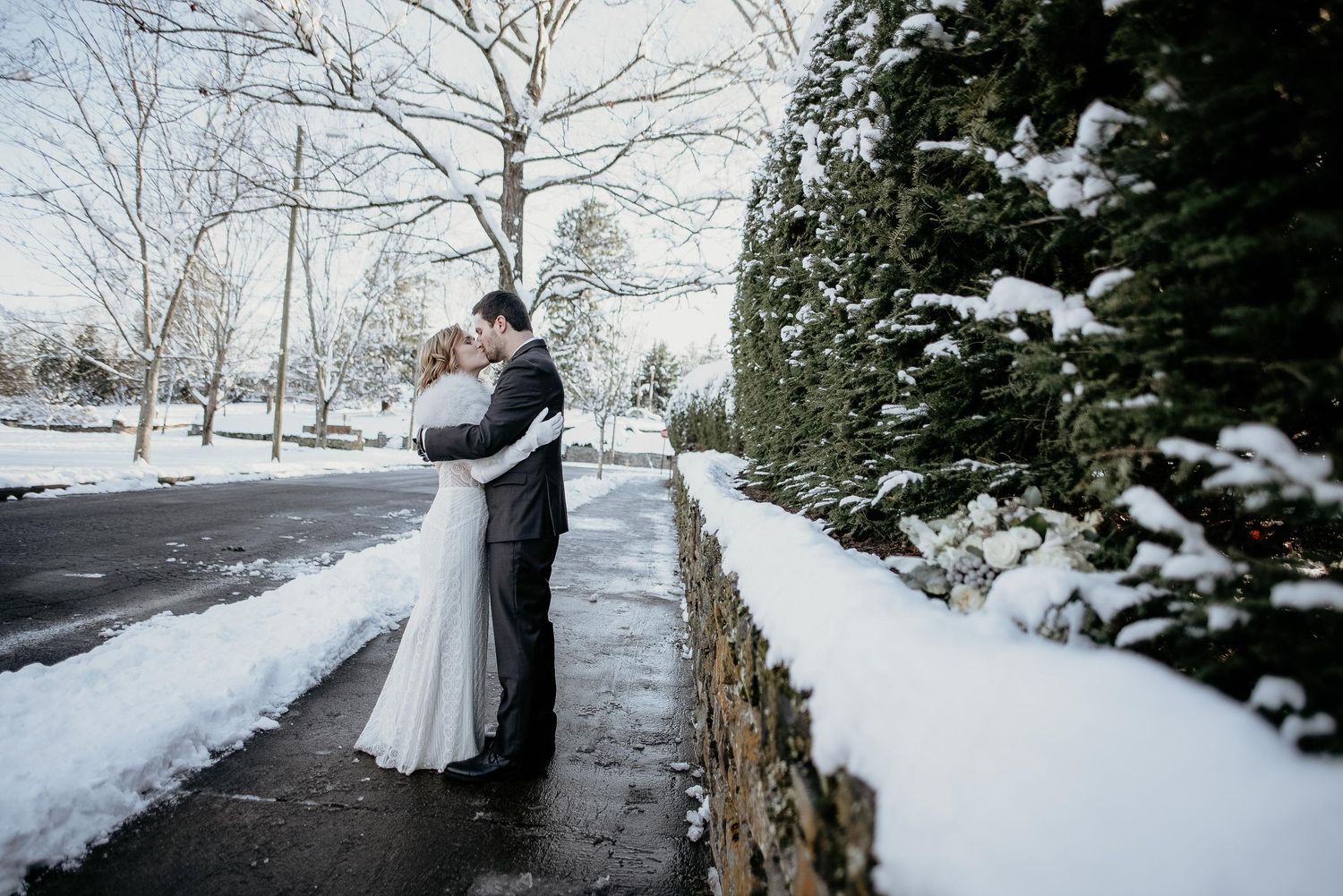 A bride and groom embrace romantically on a snowy winter day, surrounded by snow-covered trees and a stone wall.