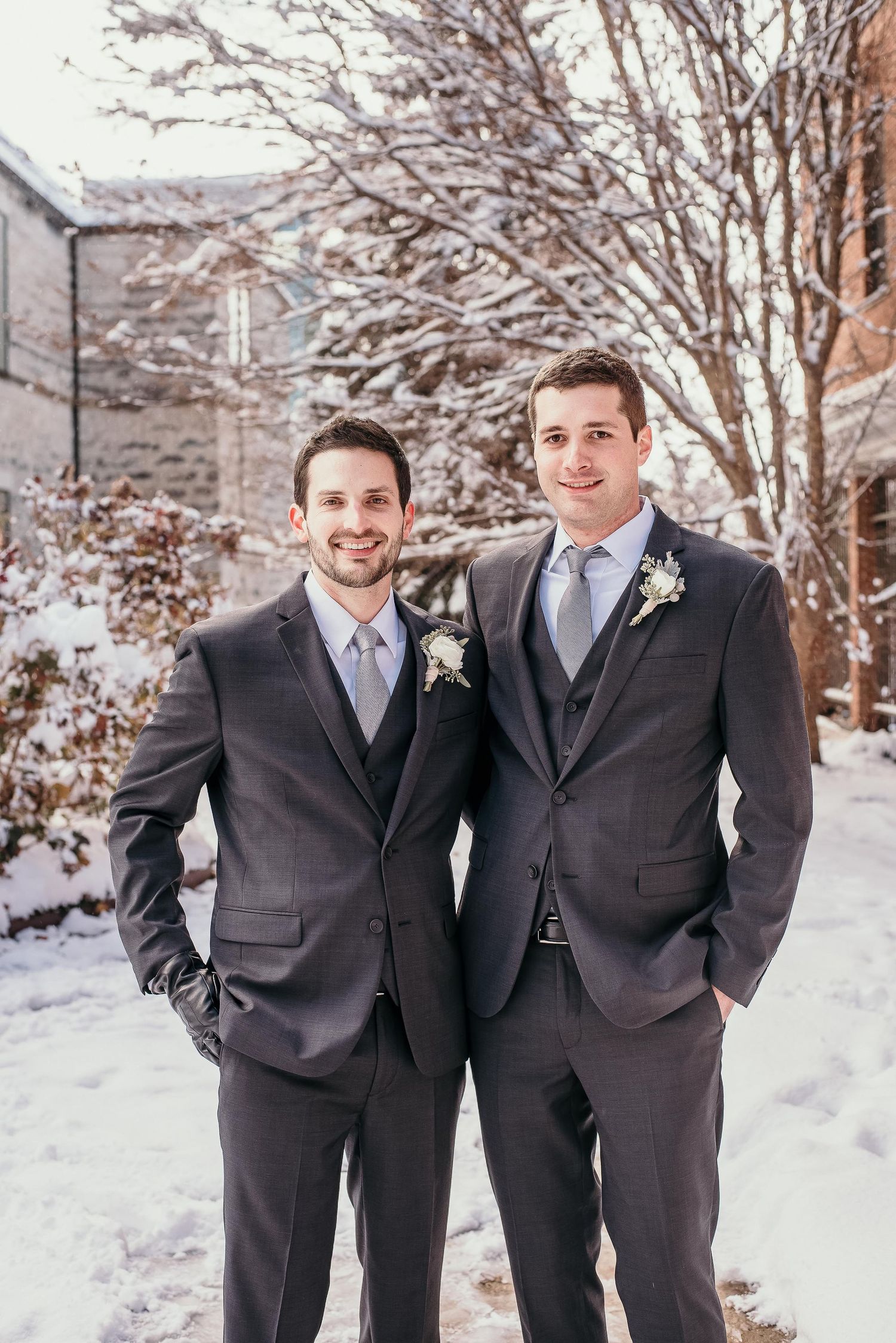 Two groomsmen in matching dark suits with boutonnieres posing outdoors in a snowy winter wedding setting.