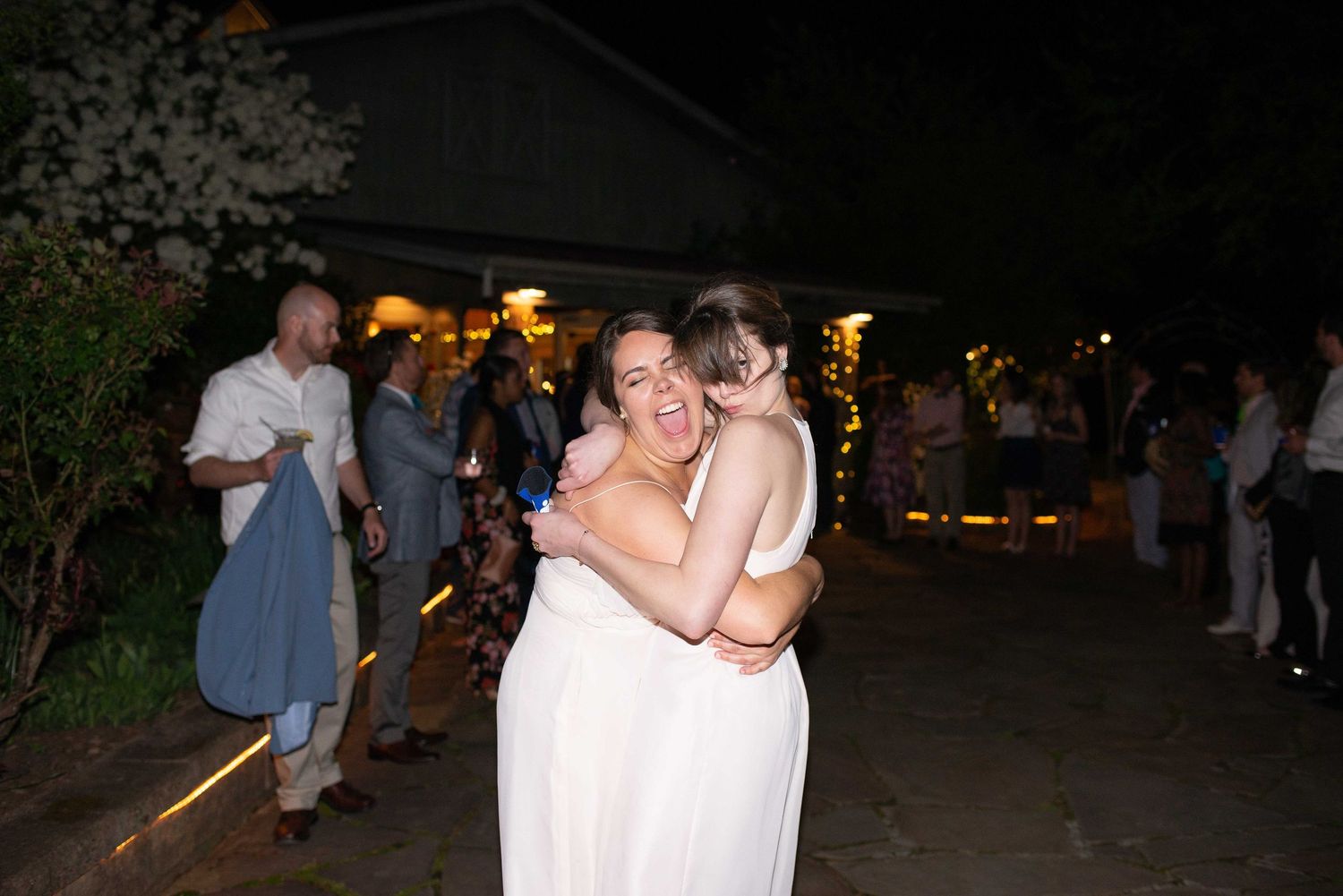 Two women in white dresses share a joyful hug at an outdoor evening wedding reception.