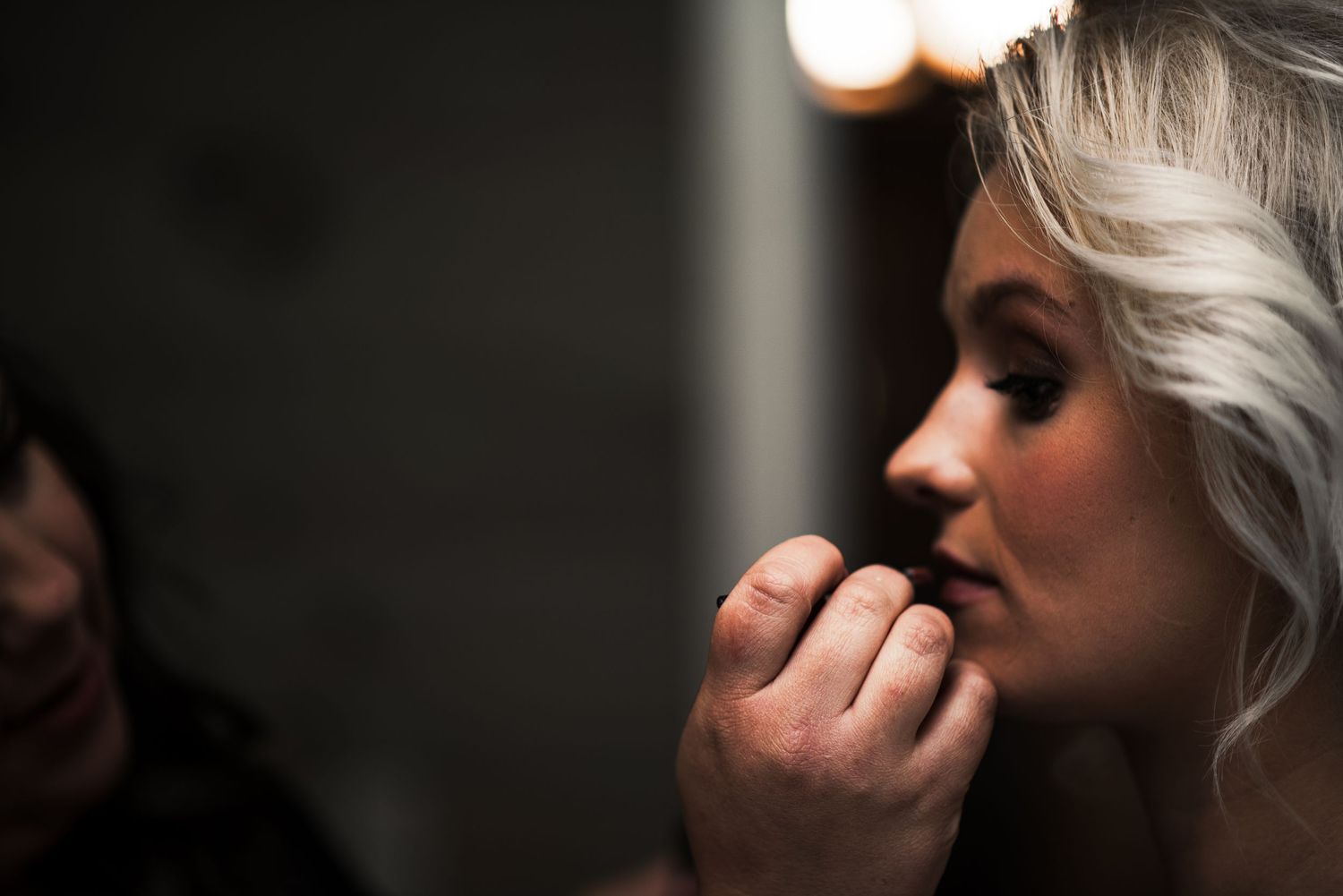 Bride having makeup applied by artist in dimly lit vanity mirror before wedding ceremony.