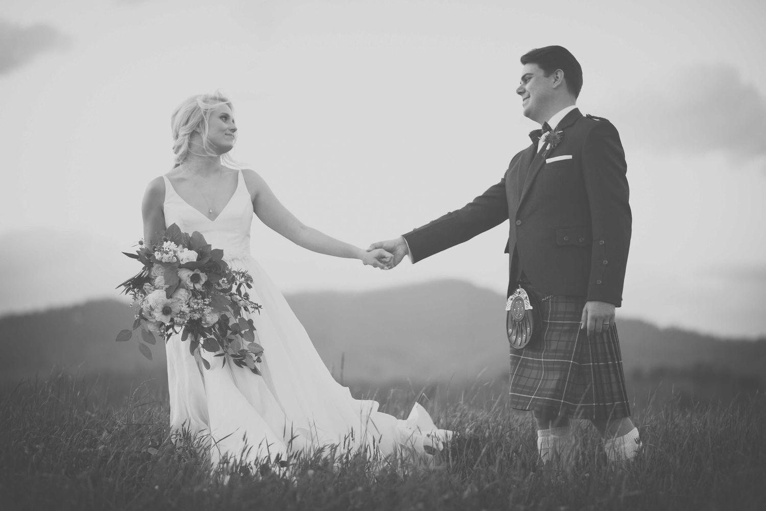 Black and white photo of a bride and groom holding hands in a field, groom wearing a traditional Scottish kilt.