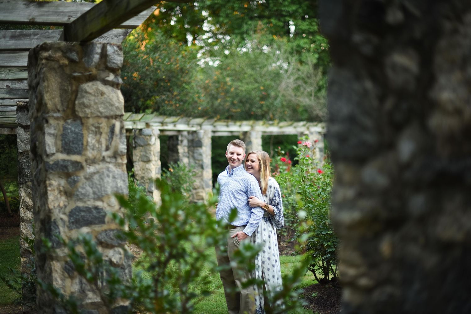 Couple embracing and smiling among stone columns in a lush garden setting.