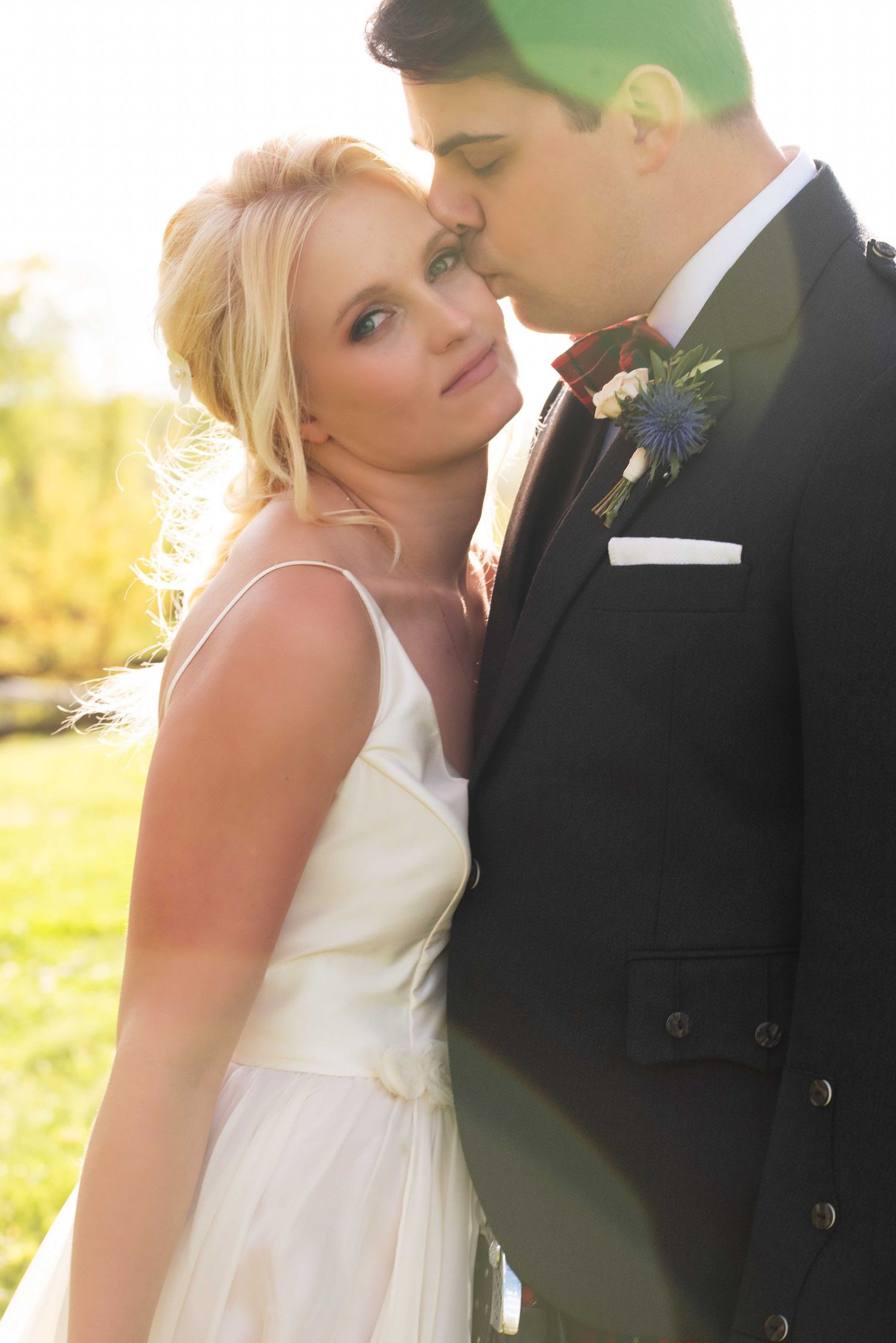 A radiant bride in a white gown leans against a groom in a dark suit with a red bow tie outdoors.