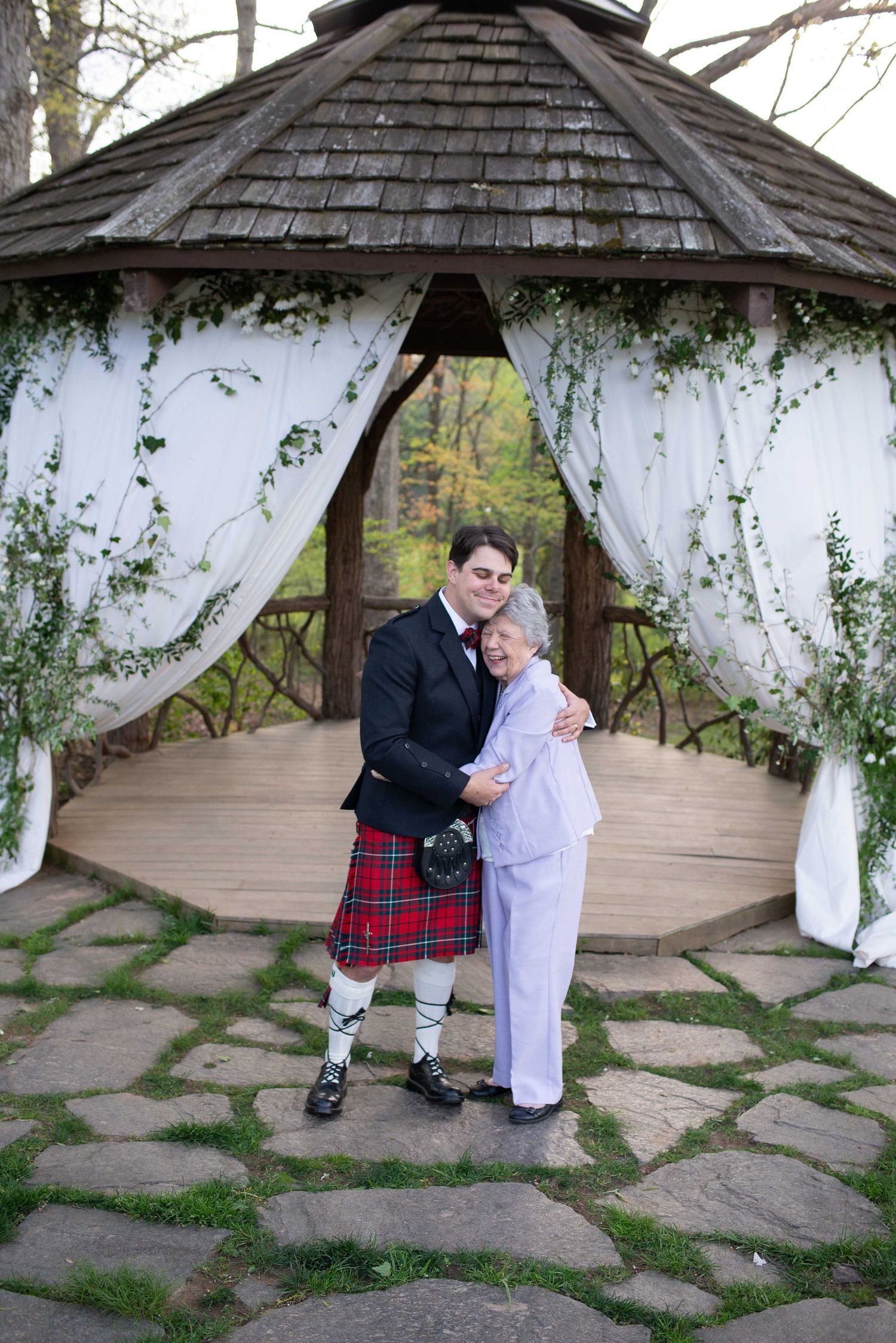 Couple embracing in a decorated gazebo, groom wearing a red tartan kilt and bride in white pantsuit on a stone path.