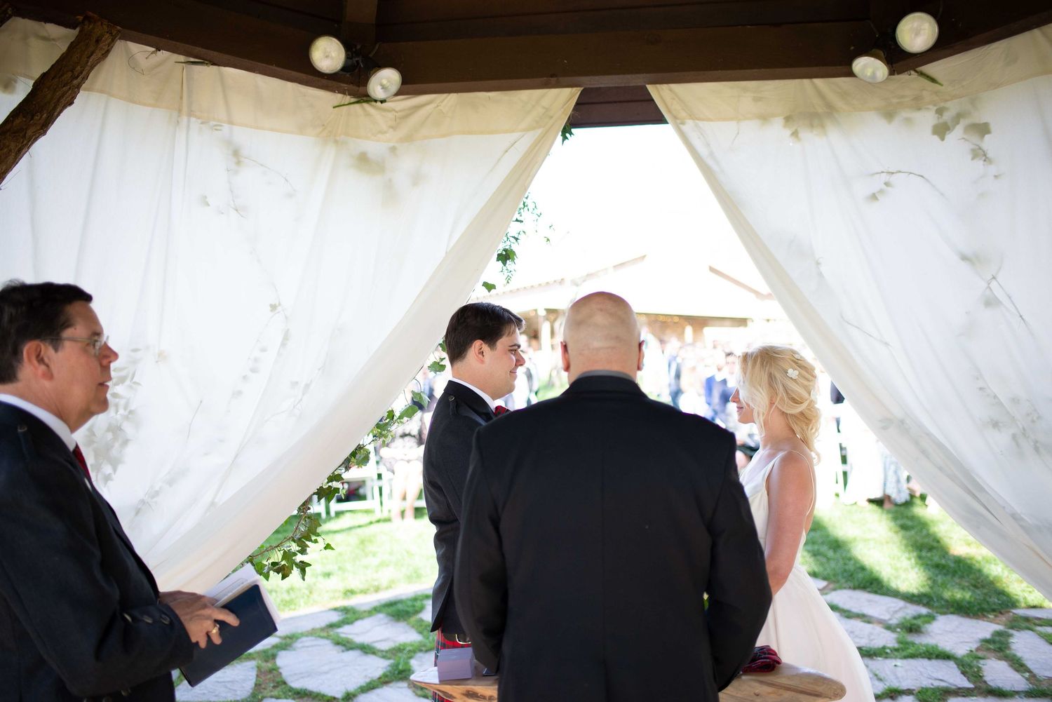 Outdoor wedding ceremony under white draped gazebo with couple exchanging vows before officiant on stone pathway.