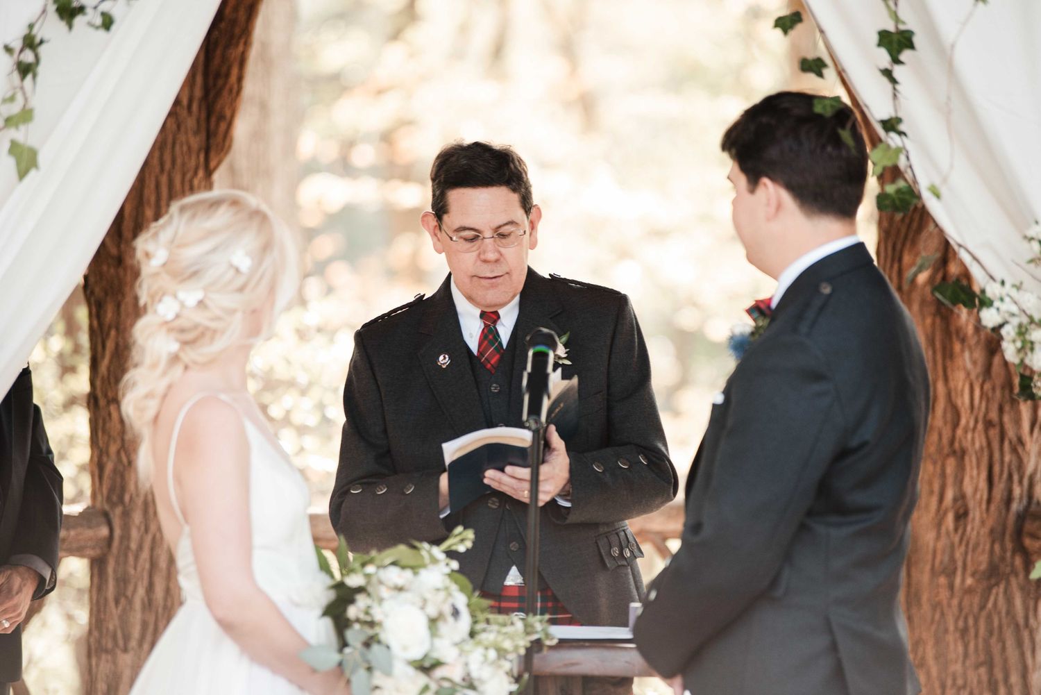 Officiant reads from book during outdoor wedding ceremony beneath floral arch with bride and groom.