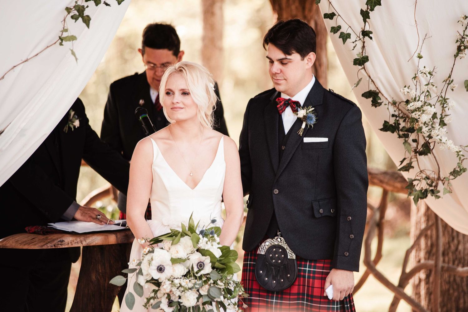 Bride in white gown and groom in red tartan kilt smile during outdoor wedding ceremony with floral backdrop.