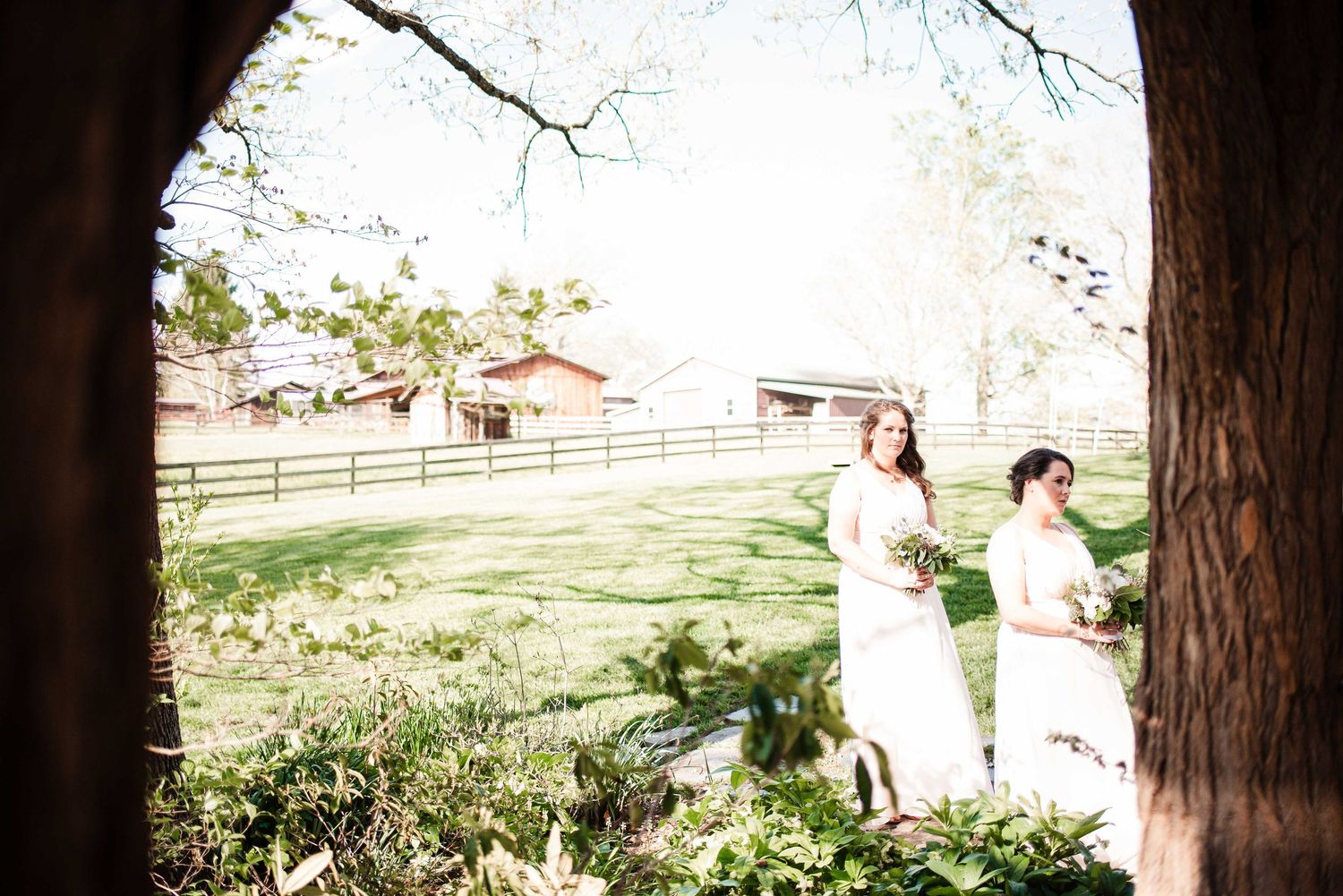 Two bridesmaids in white dresses holding bouquets stand on lush green lawn near rustic barn and wooden fence.