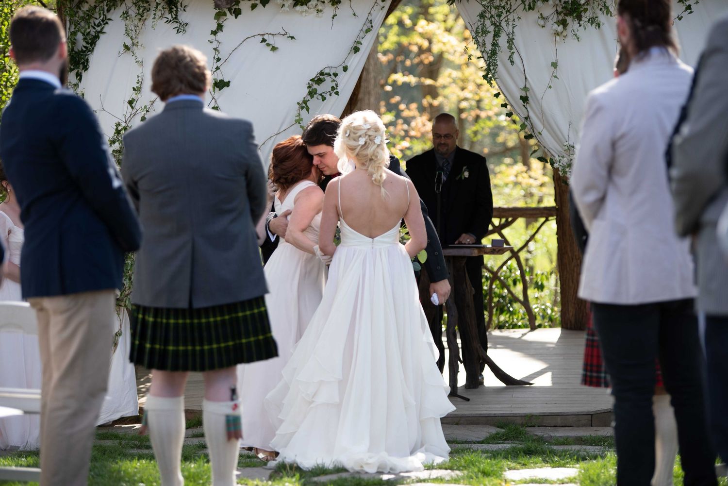 Outdoor wedding ceremony with bride in white gown, guests in kilts, under floral arch on sunny day.