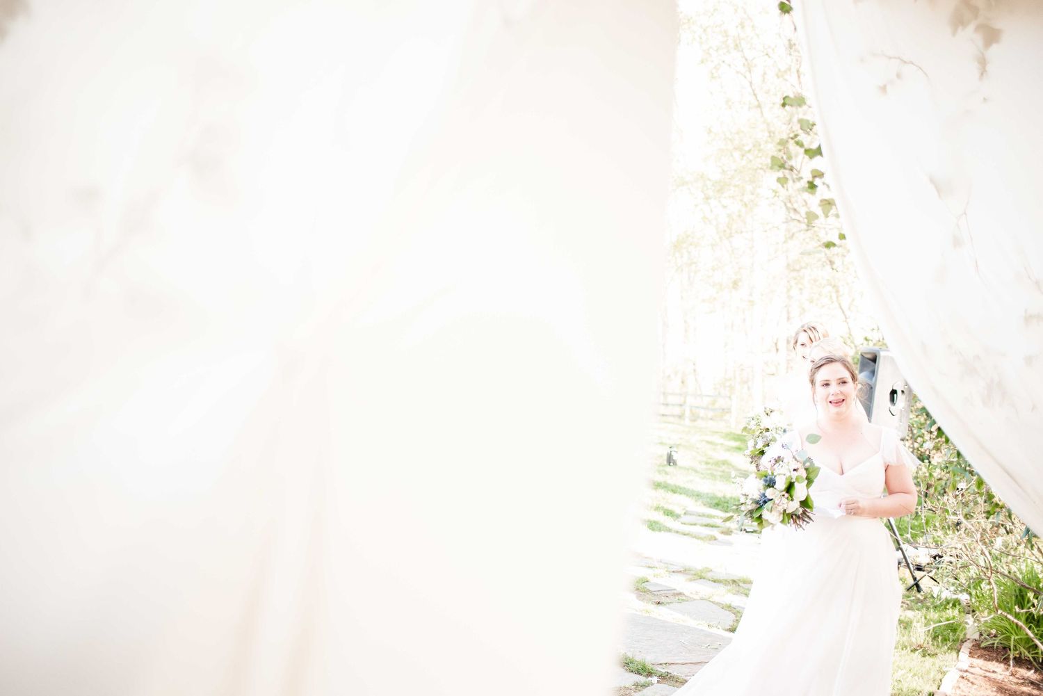 Elegant bride in white lace gown holding bouquet, standing between draped fabric panels in a sunlit outdoor setting.