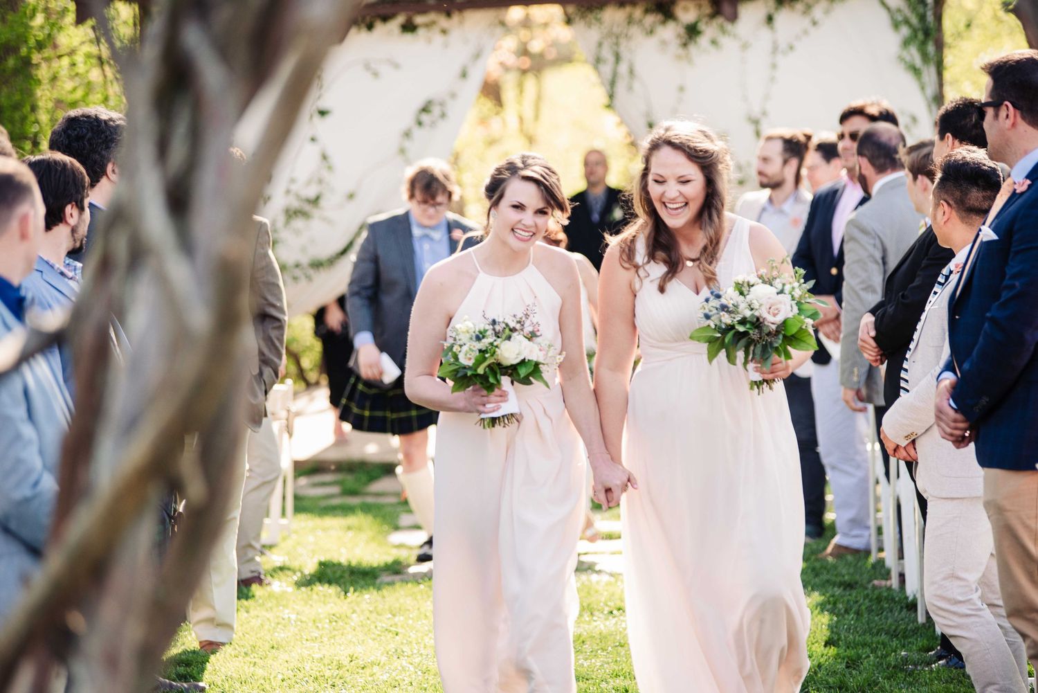 Two smiling bridesmaids in blush pink gowns walk down outdoor wedding aisle holding white floral bouquets.