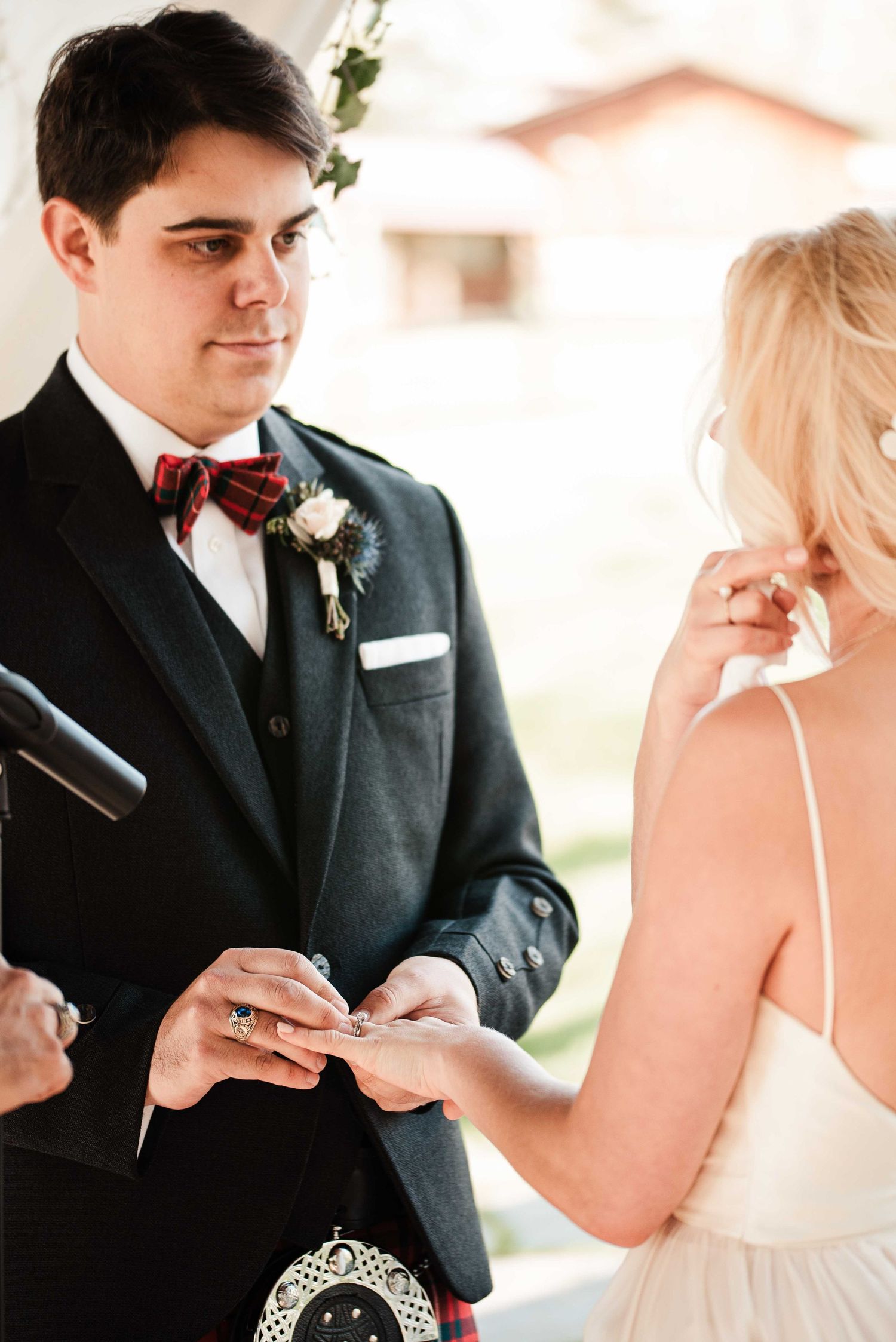 Groom in dark suit with tartan kilt and red plaid bow tie exchanges wedding rings with blonde bride during ceremony.