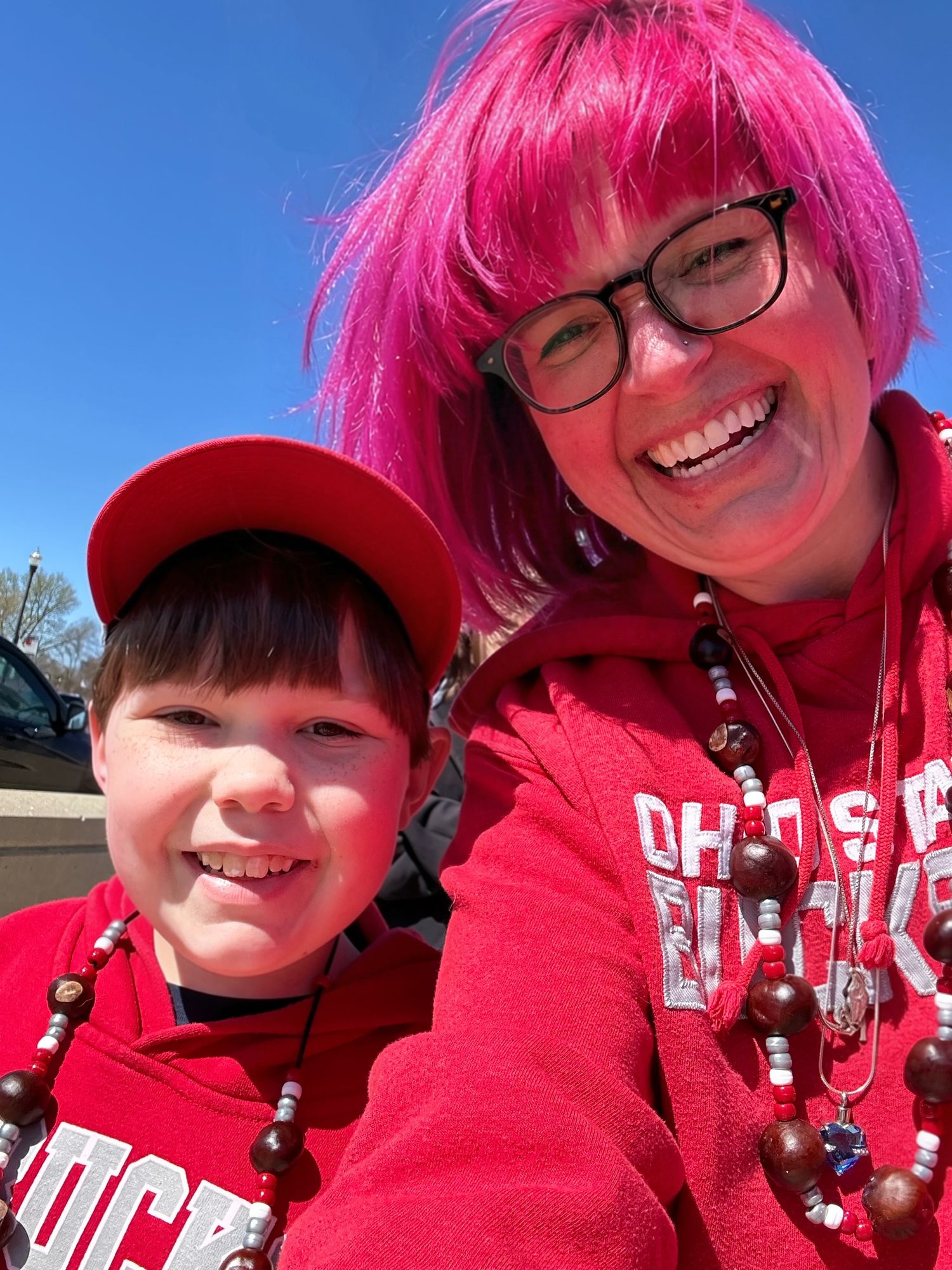 Woman with pink hair and glasses smiles with young boy, both wearing red Ohio State Buckeyes gear outdoors.
