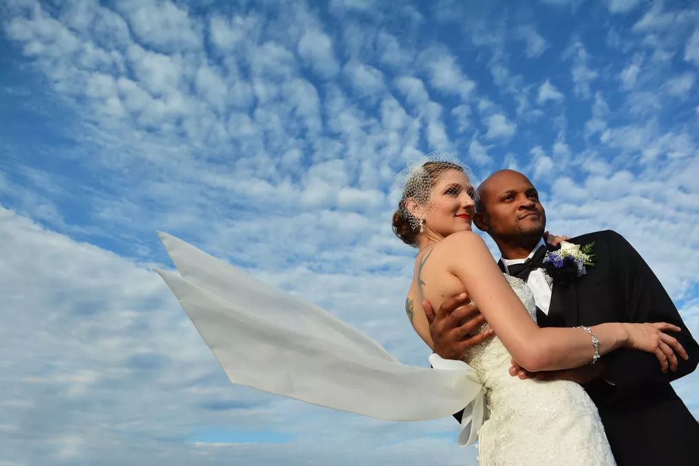 Bride and groom embrace outdoors with veil flowing in wind against dramatic blue sky with white clouds on wedding day.