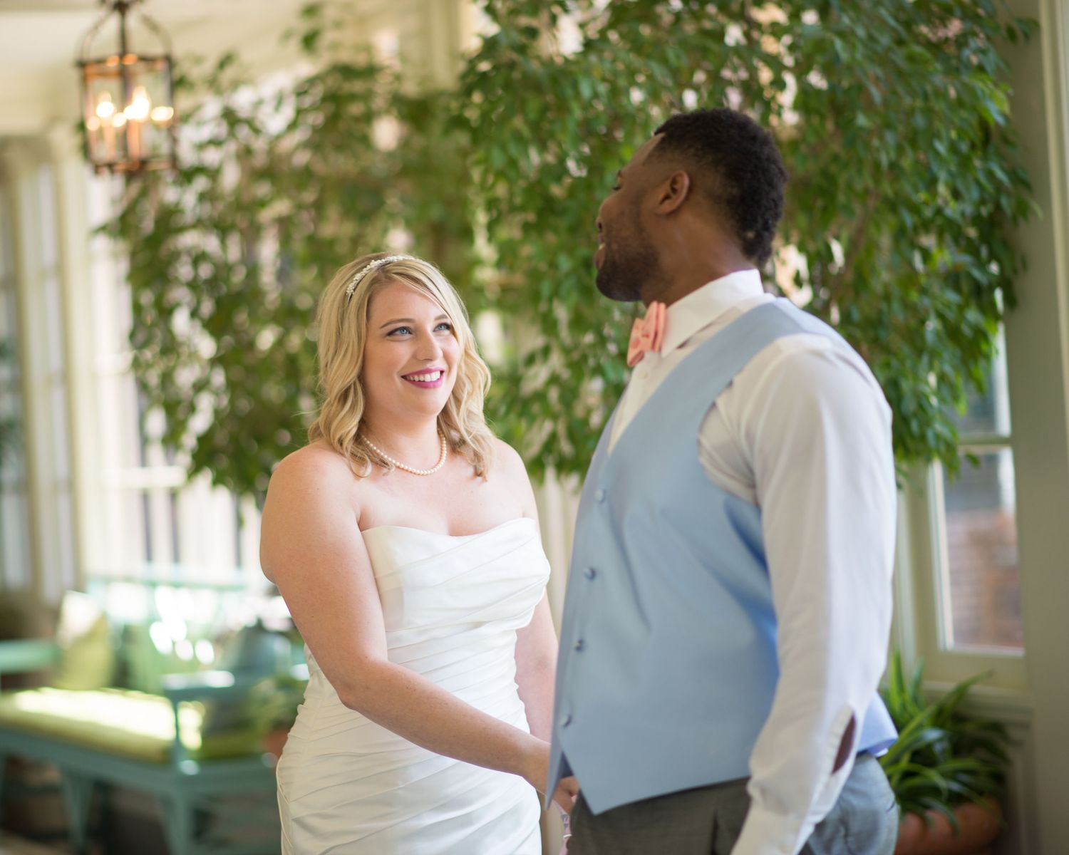 Interracial couple holding hands and smiling at each other during wedding ceremony indoors with lush green plants.
