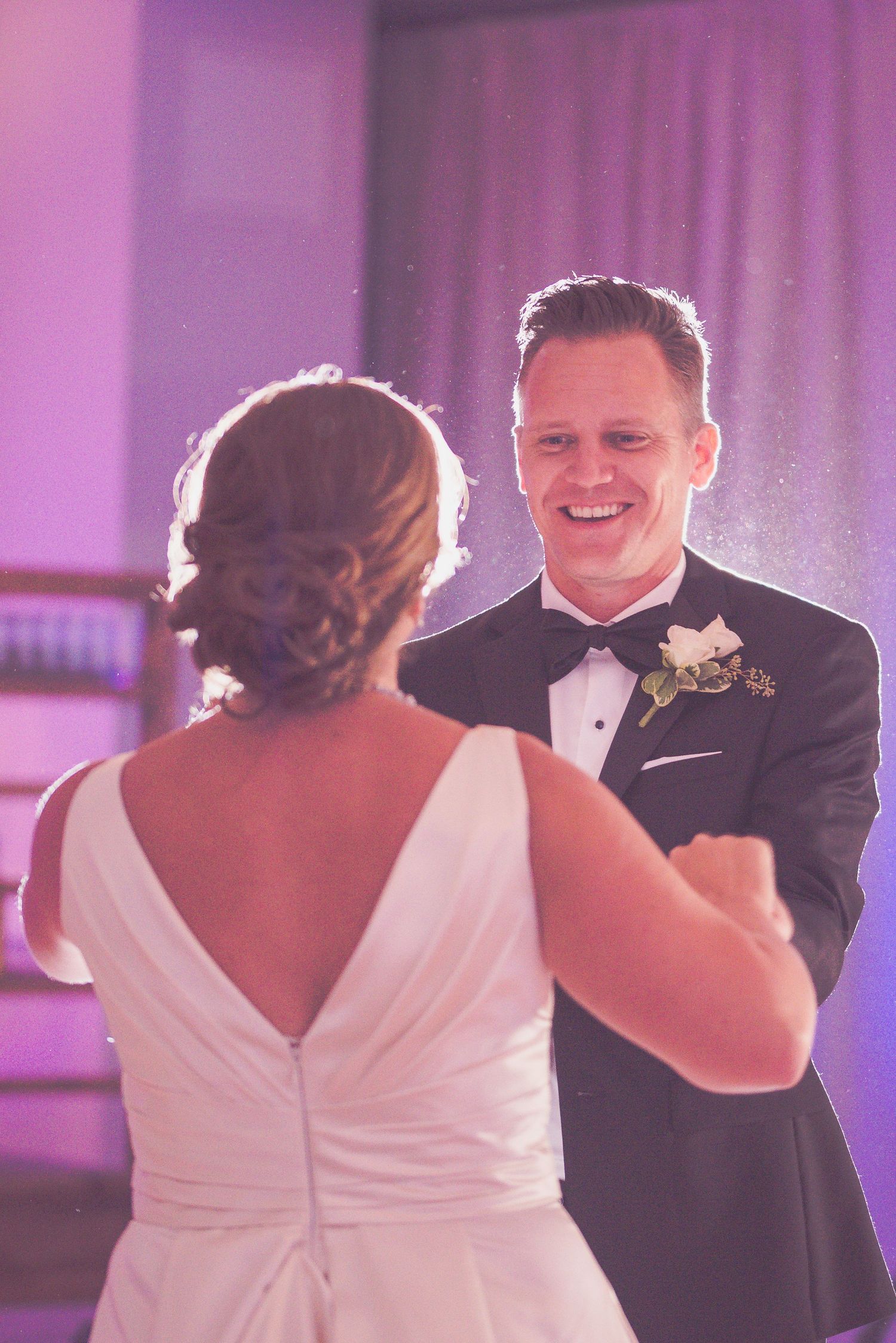 Groom smiles lovingly at bride during their first dance under soft purple uplighting at wedding reception.